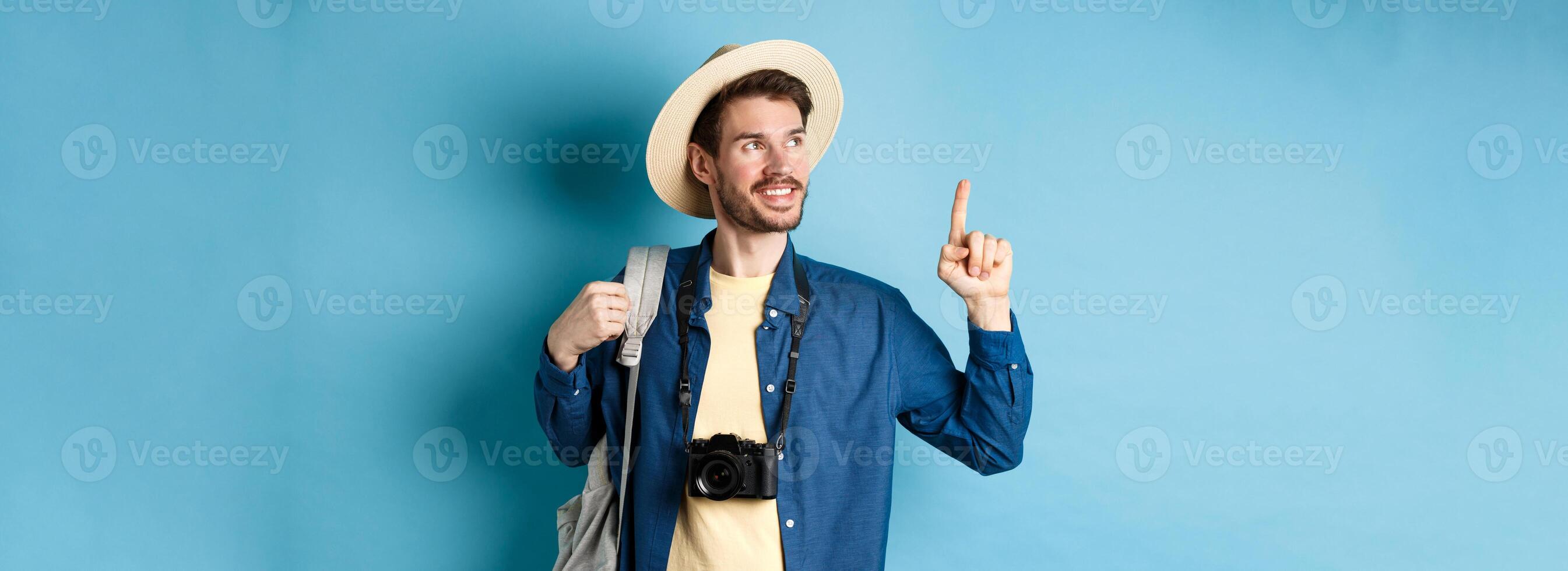 Handsome positive guy on vacation, wearing summer hat, looking and pointing up at logo, smiling pleased, standing with camera and backpack on blue background photo
