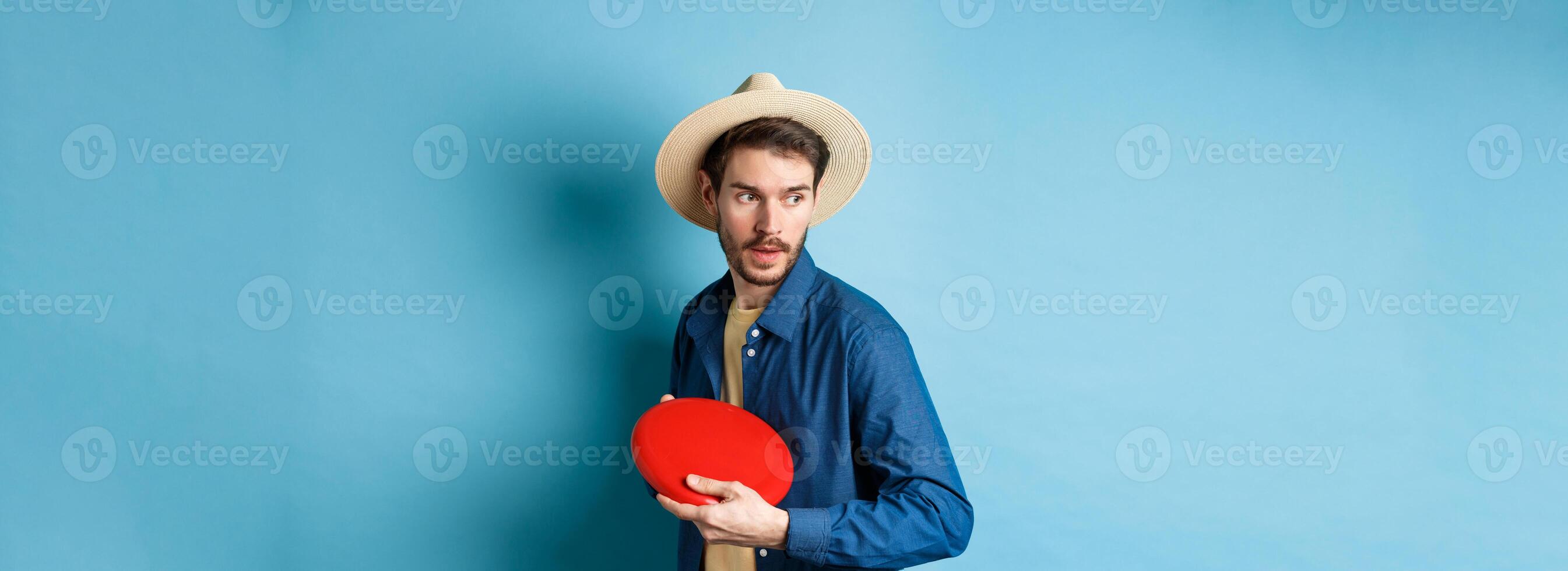 Handsome young guy in summer hat playing frisbee, throwing it aside, standing on blue background photo