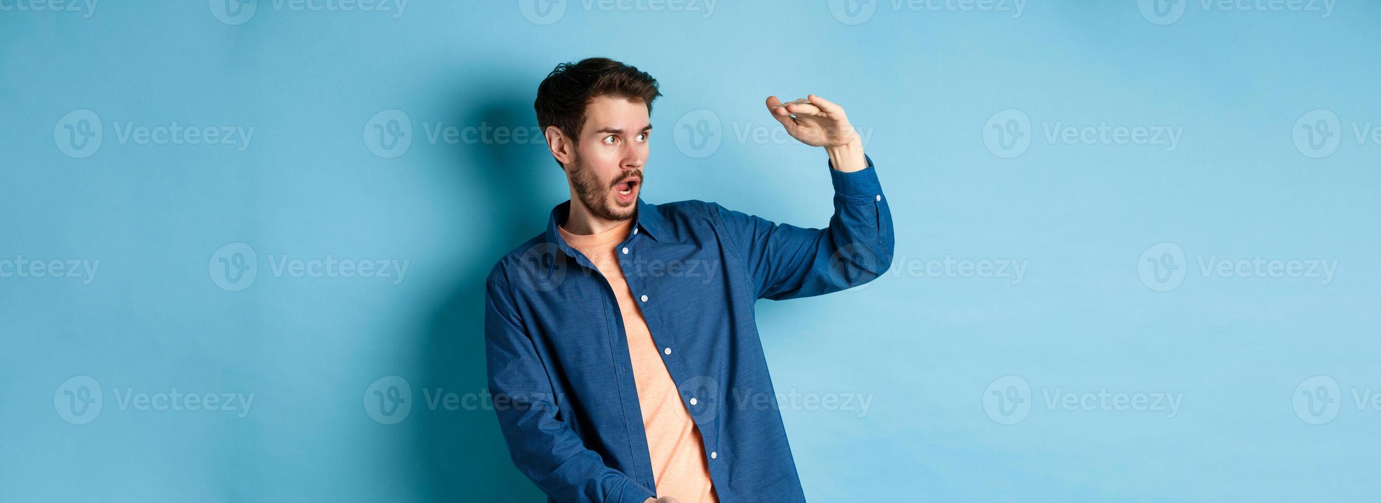 Impressed caucasian guy staring at empty space with big object, shaping large size, standing on blue background photo