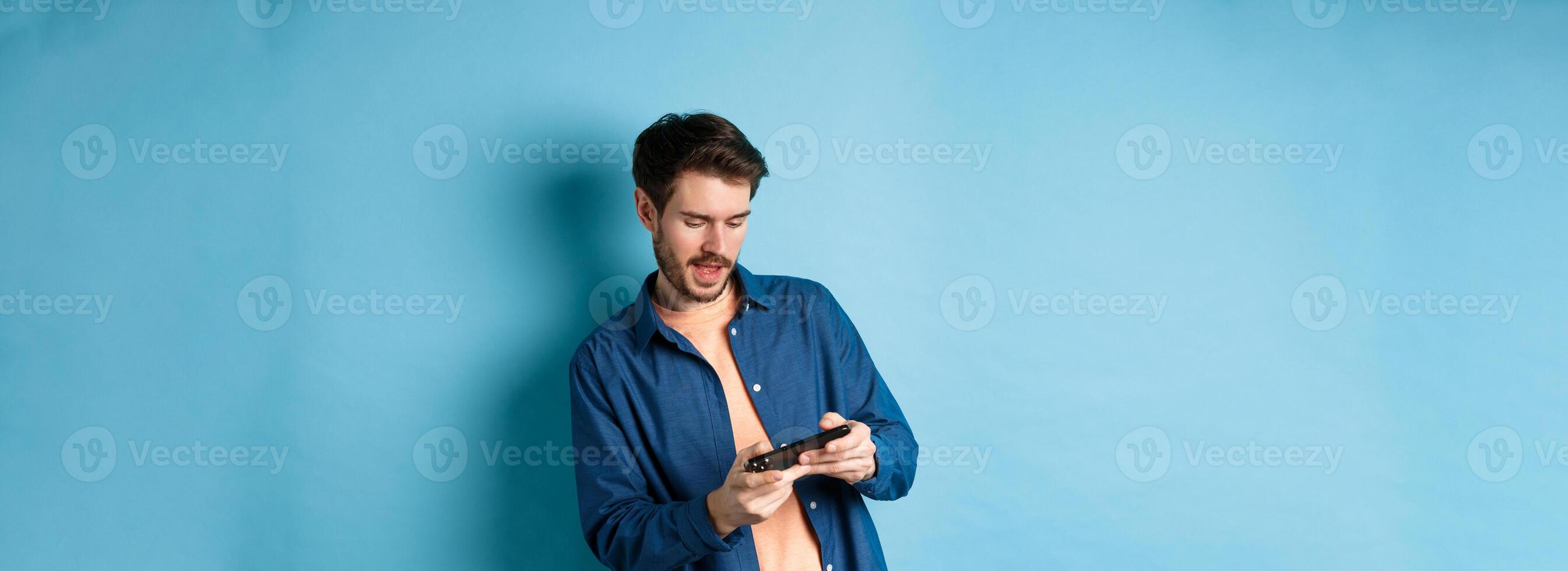 Young guy playing game on mobile phone, tilt body and holding smartphone horizontally, standing on blue background photo