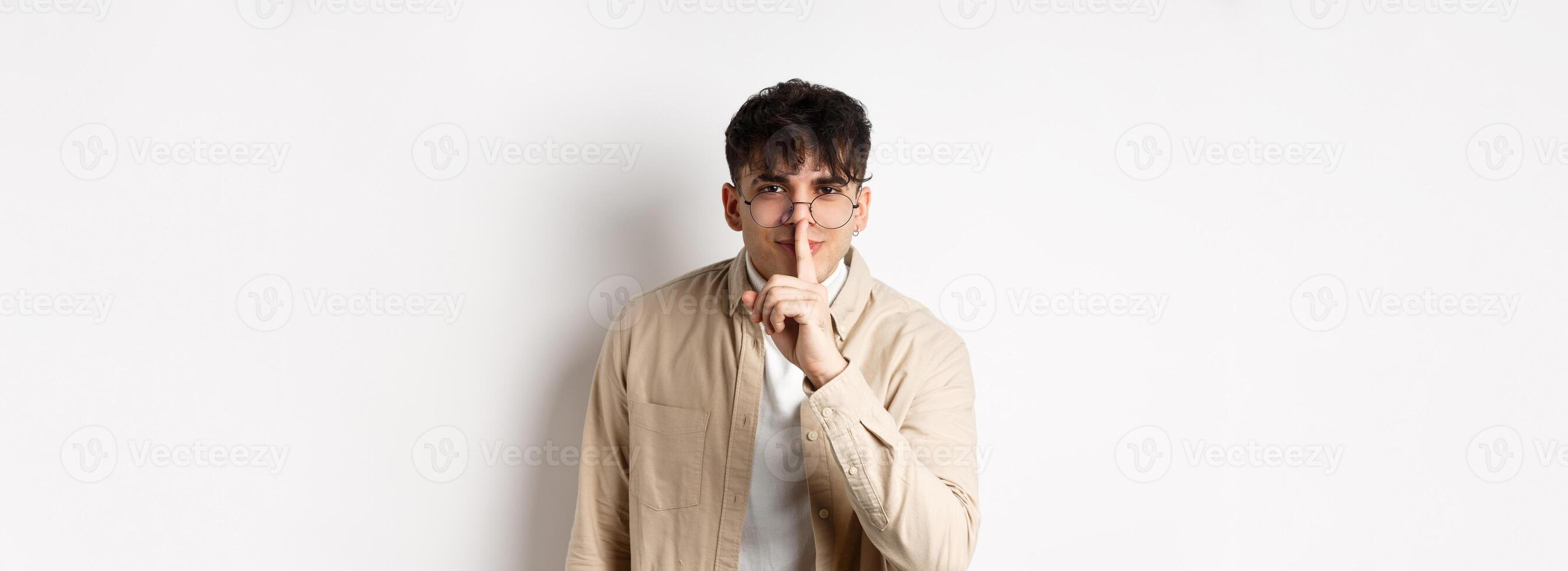 Cheerful handsome man in glasses share a secret, making surprise, lean at camera with hush gesture and gentle smile, telling to be quiet, show taboo sign, white background photo