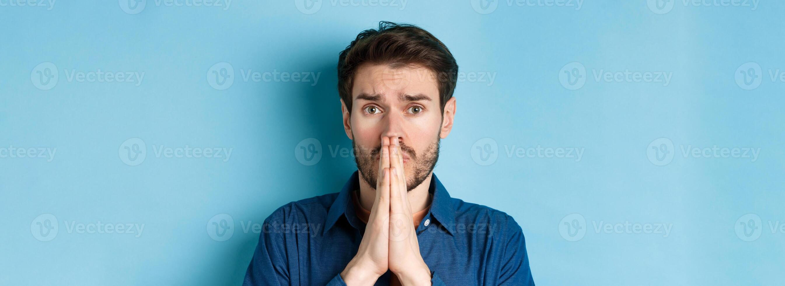 Close up of sad young man pleading, begging for help and say please, standing on blue background photo