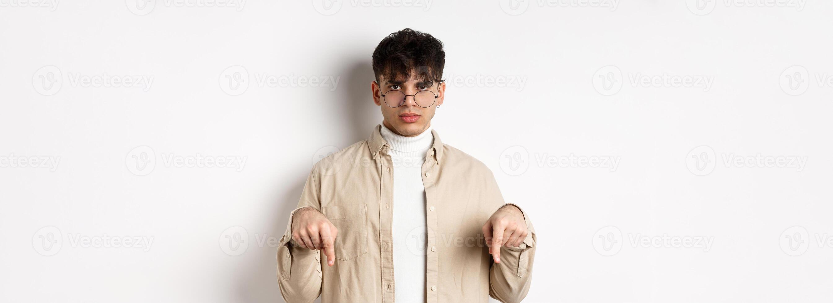 Portrait of handsome hipster guy in glasses showing an ad, pointing fingers down at logo, standing on white background photo