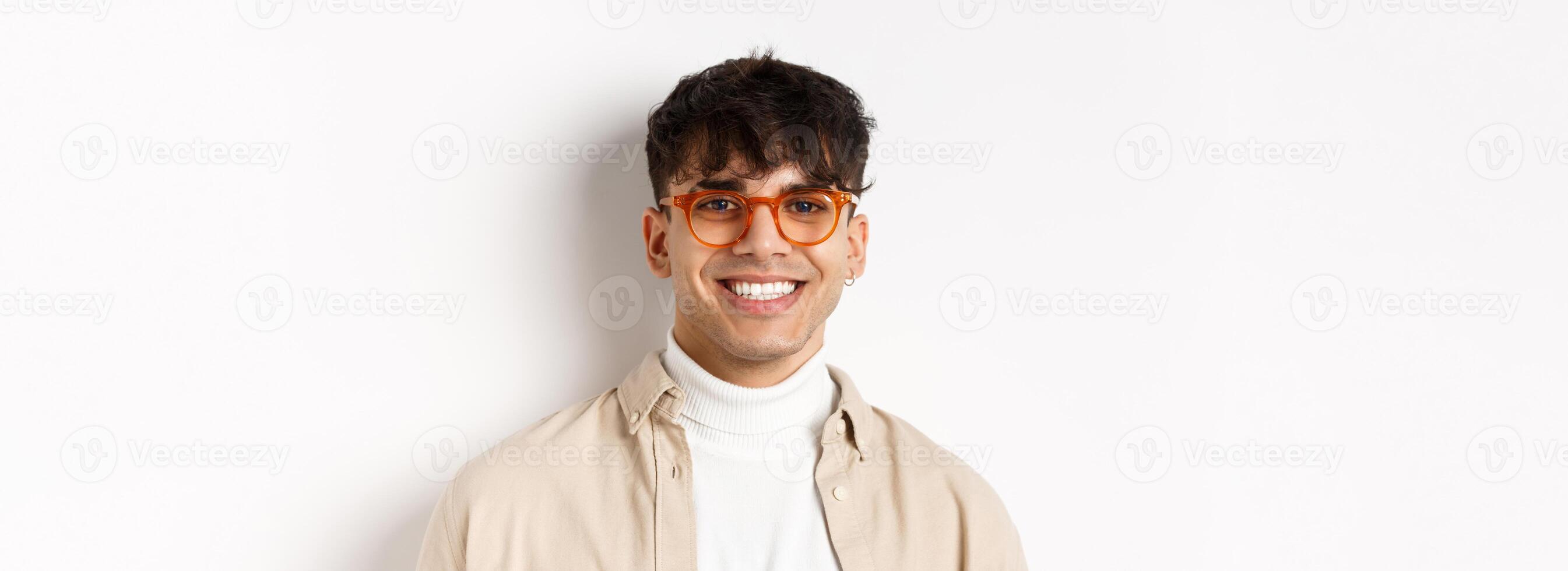 Close-up of handsome hipster guy with natural smile, wearing glasses and earrings, looking happy at camera, standing on white background photo