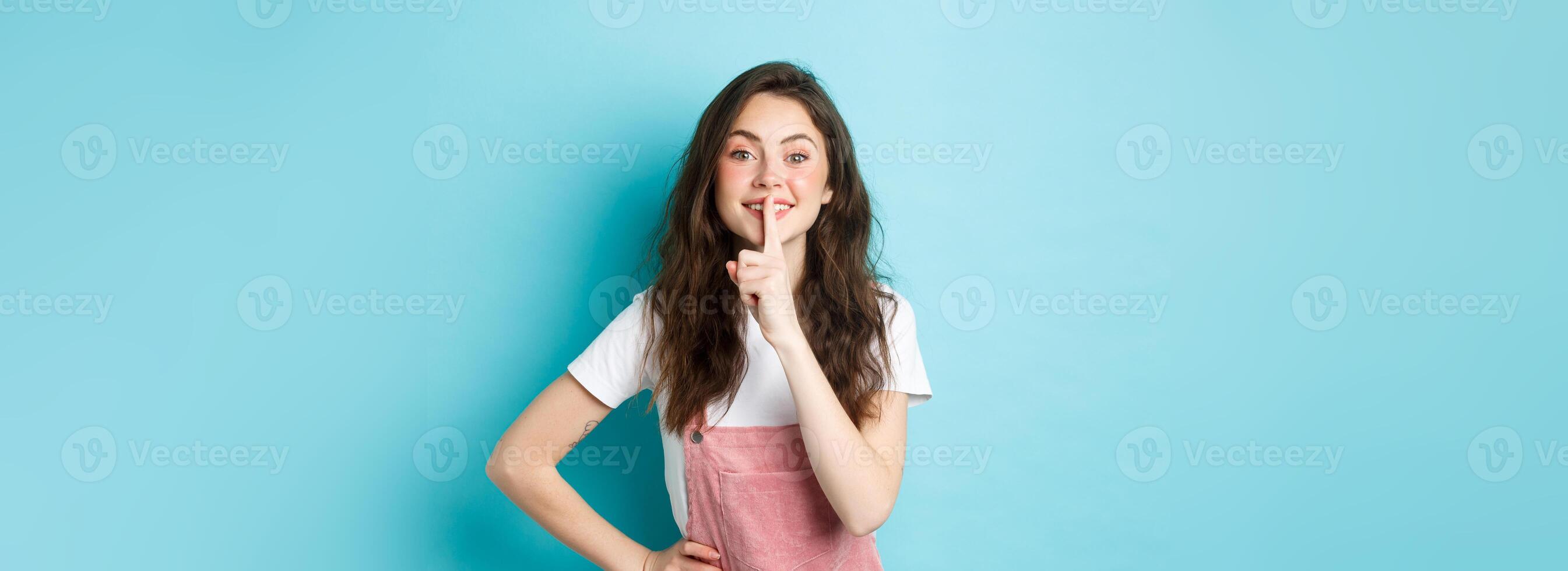 Shh be quiet. Cute brunette girl shushing and smiling, having a secret, holding finger on mouth and looking kind at camera, standing over blue background photo