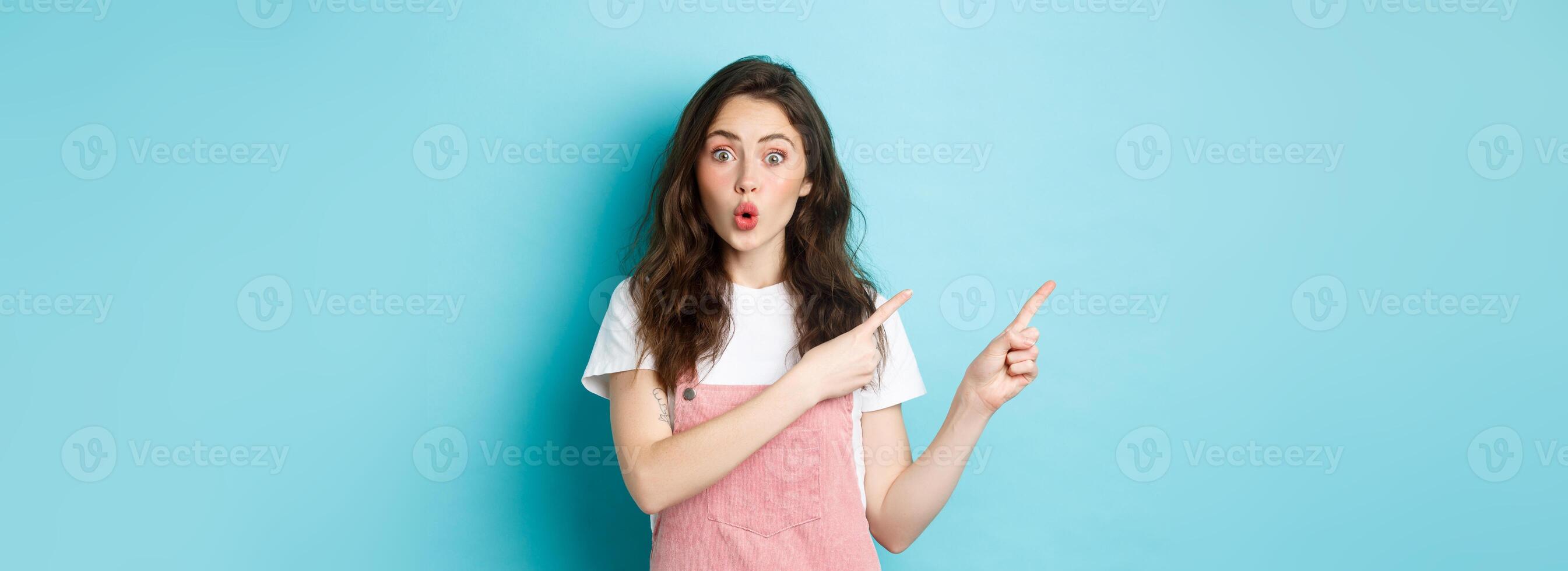 Wow check this out. Amazed brunette female model pucker lips and look intrigued, pointing fingers left at something interesting, showing banner, blue background photo