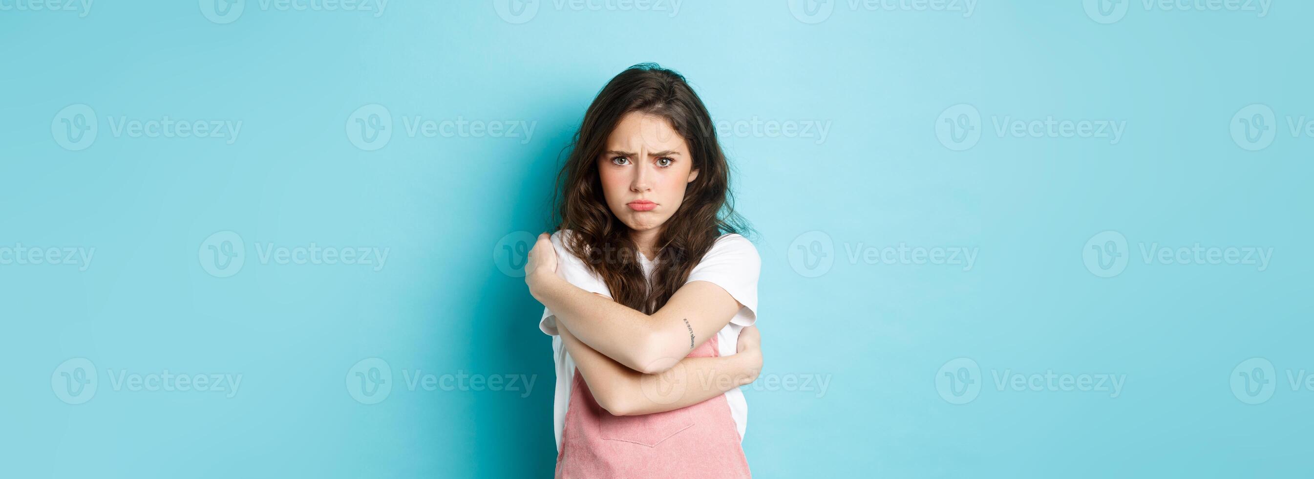 Sad and gloomy young woman pouting, comforting herself with cuddles, hugging her body and looking upset about something unfair, standing against blue background photo