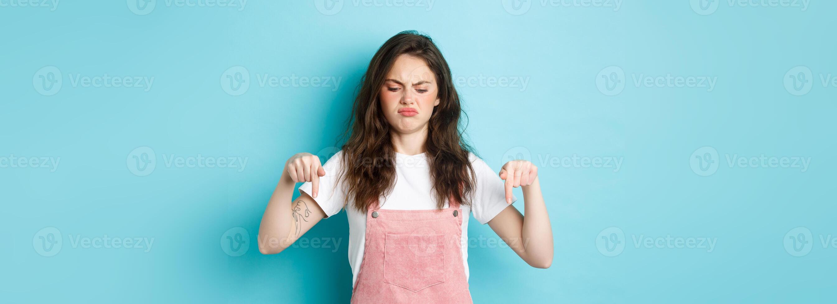 Sounds bad. Displeased grumpy brunette girl frowning and sulking moody, pointing fingers down and look disappointed, standing against blue background photo