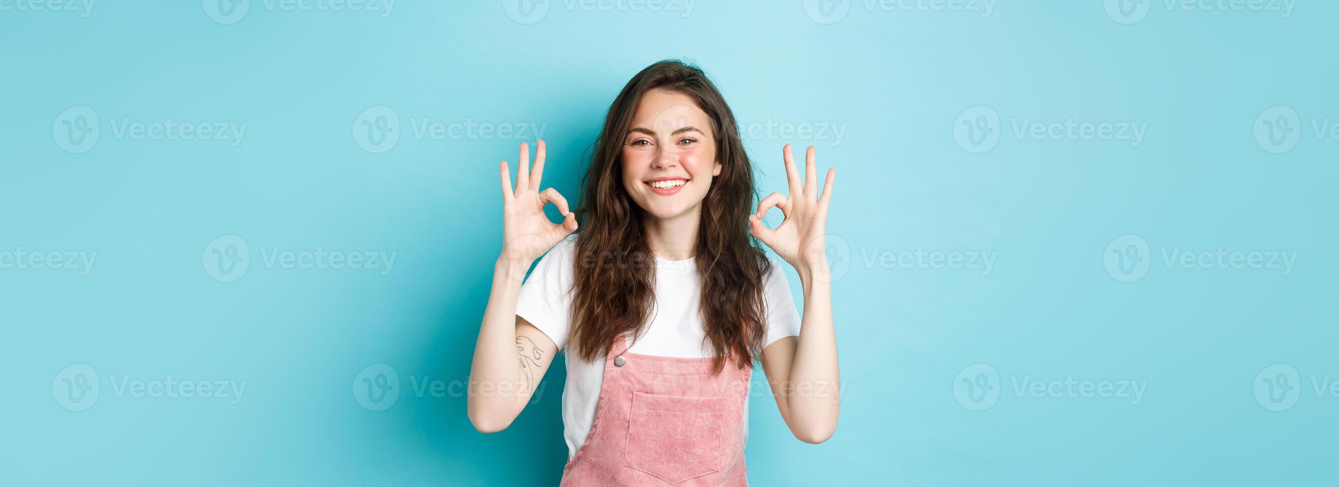 Image of smiling brunette female model say yes, showing okay signs in approval, agree or praise good choice, recommending product, standing against blue background photo
