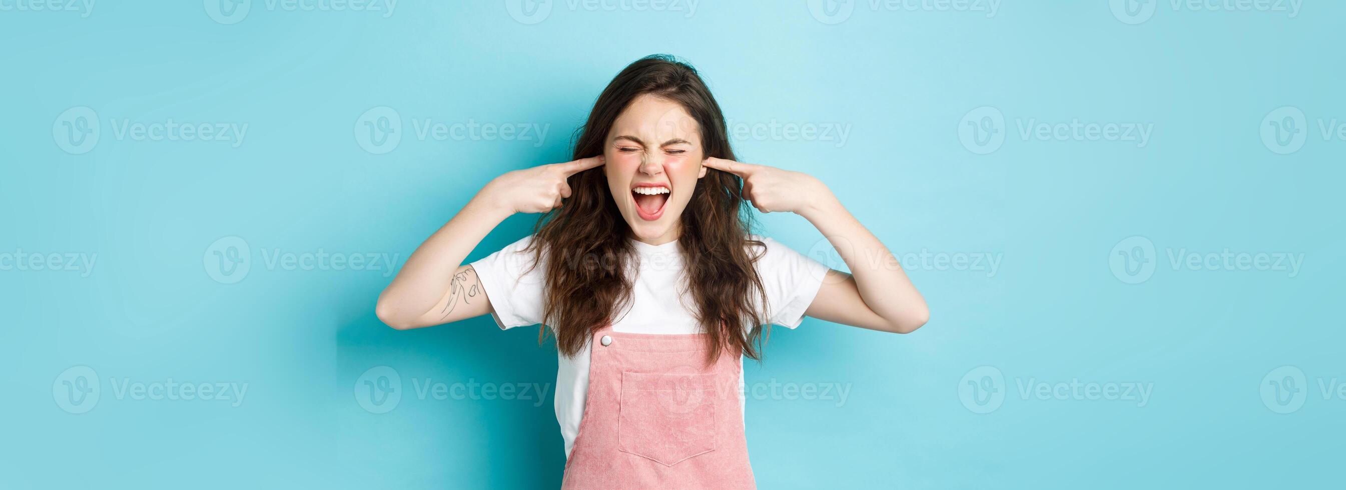 Annoyed young brunette woman screaming and shutting ears, blocking annoying sound, ignoring someone, hear awful noise, standing against blue background photo