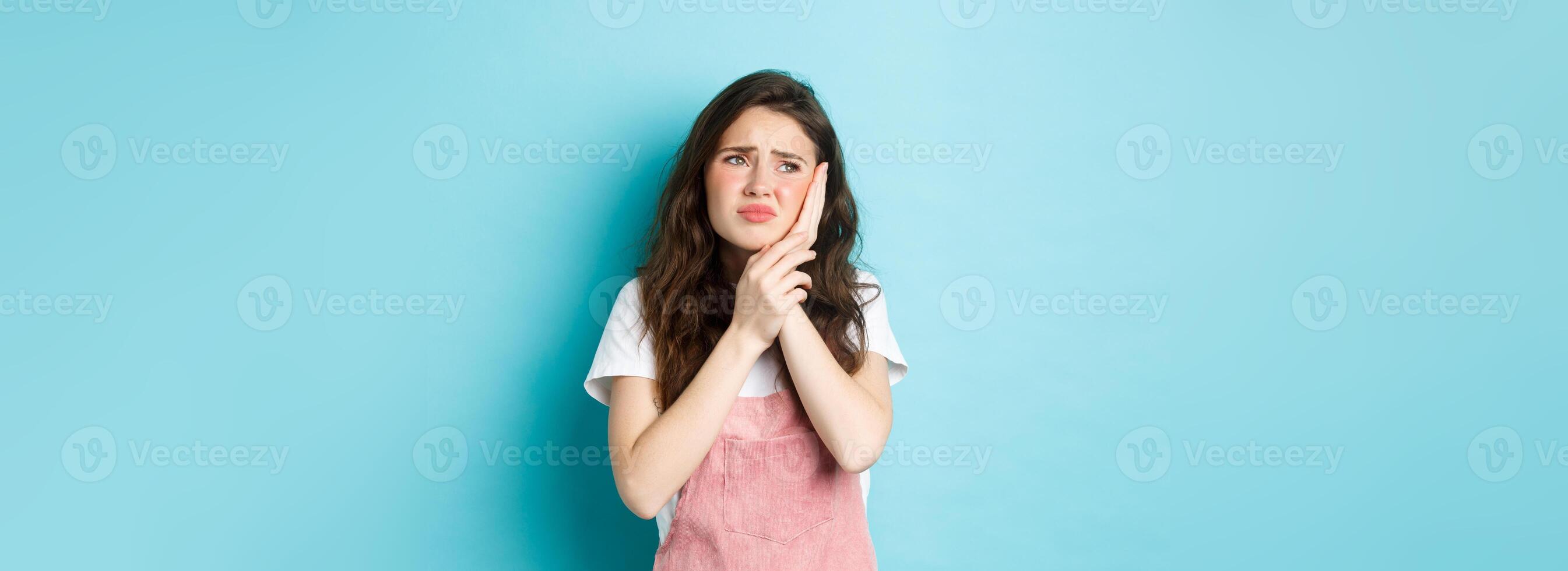 Portrait of frowning girl touching cheek, having toothache, looking at upper left corner stomatology clinic banner, suffering tooth decay, standing against blue background photo
