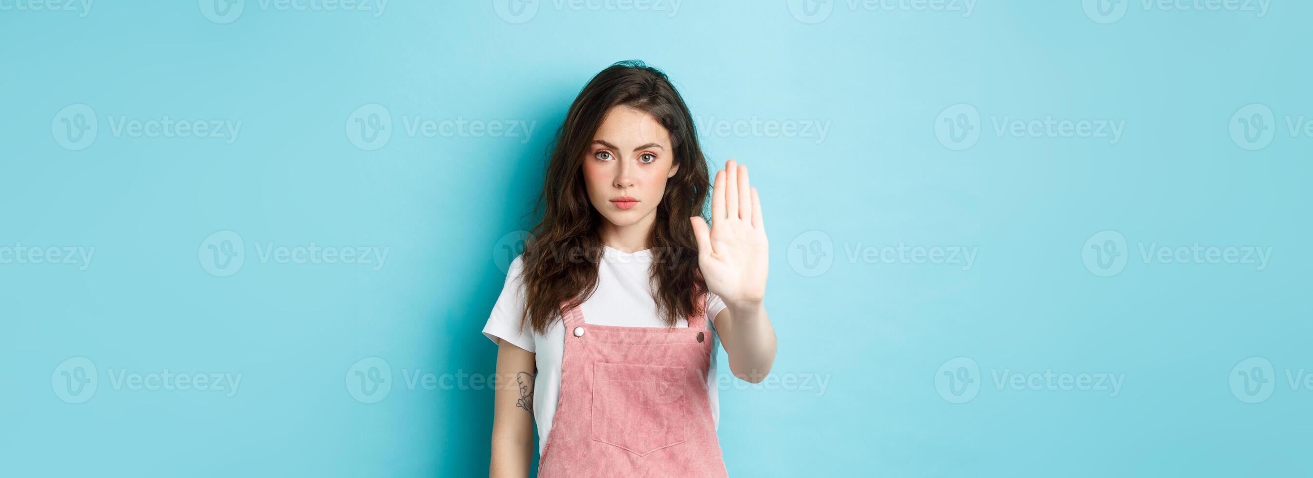 Young brunette woman with curly hairstyle, raising hand in block gesture, say stop or no, refuse bad offer, rejecting something, standing against blue background photo