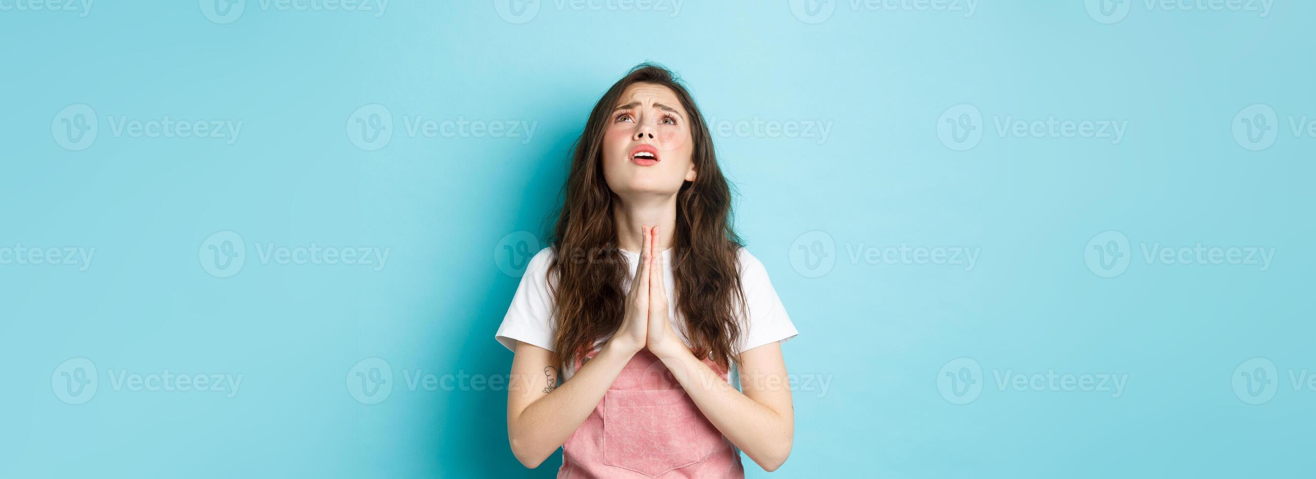 Desperate young brunette woman begging God, holding hands in pray and looking up at sky with hopeful face, pleading for dream come true, standing against blue background photo
