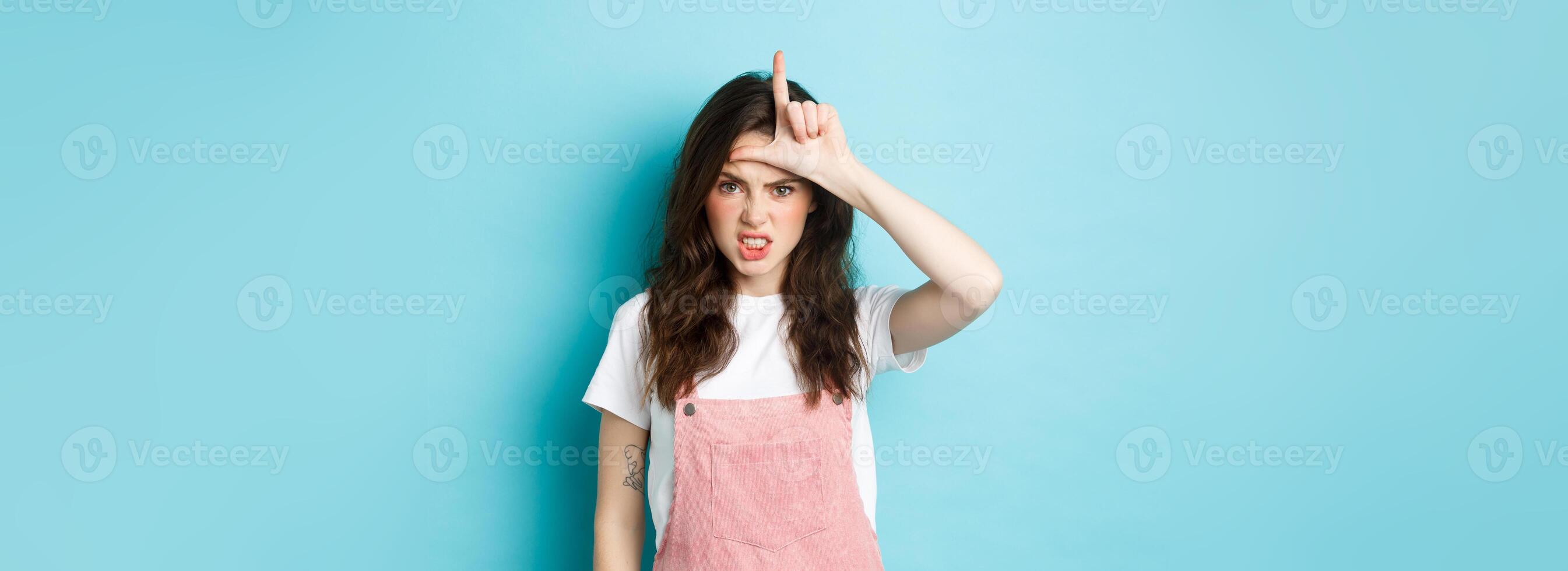 Sassy young woman despise her rival, showing loser gesture on forehead, making letter L with hand and staring with dismay at camera, standing against blue background photo