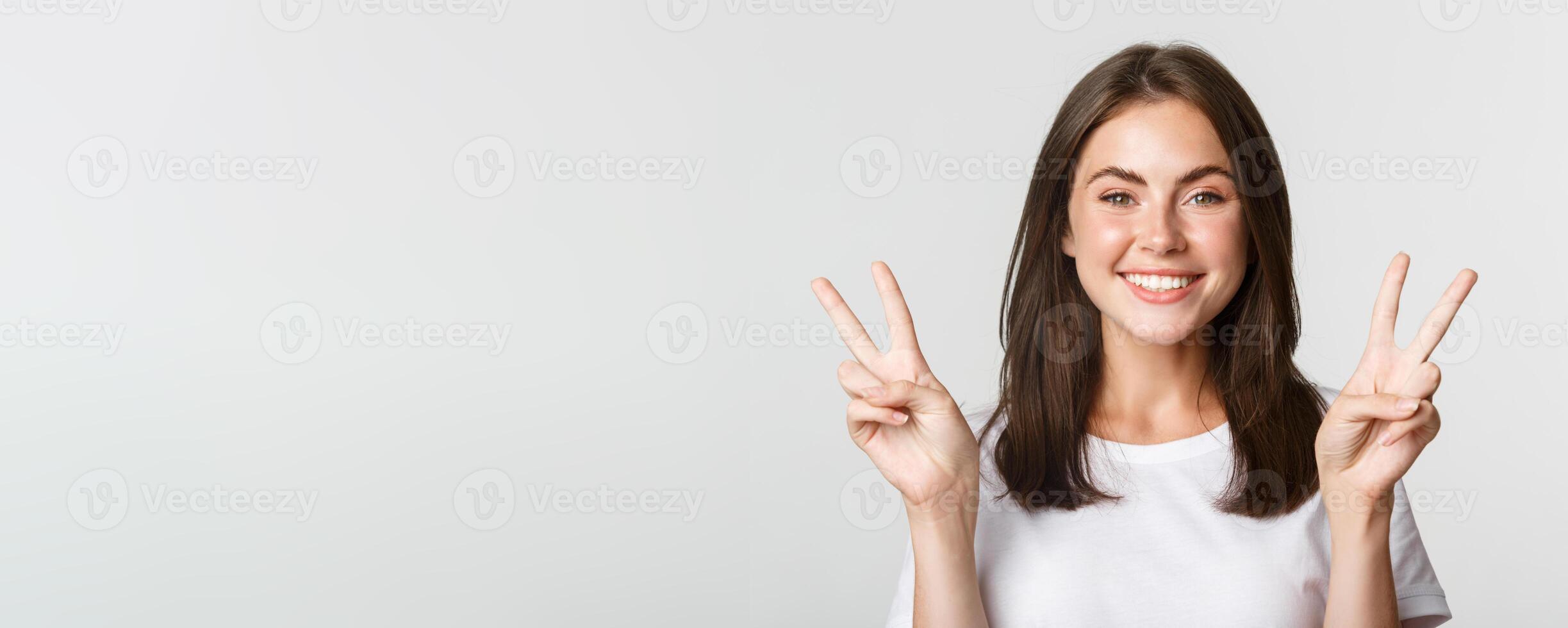 Portrait of beautiful and happy brunette girl smiling and showing peace gestures, standing white background photo