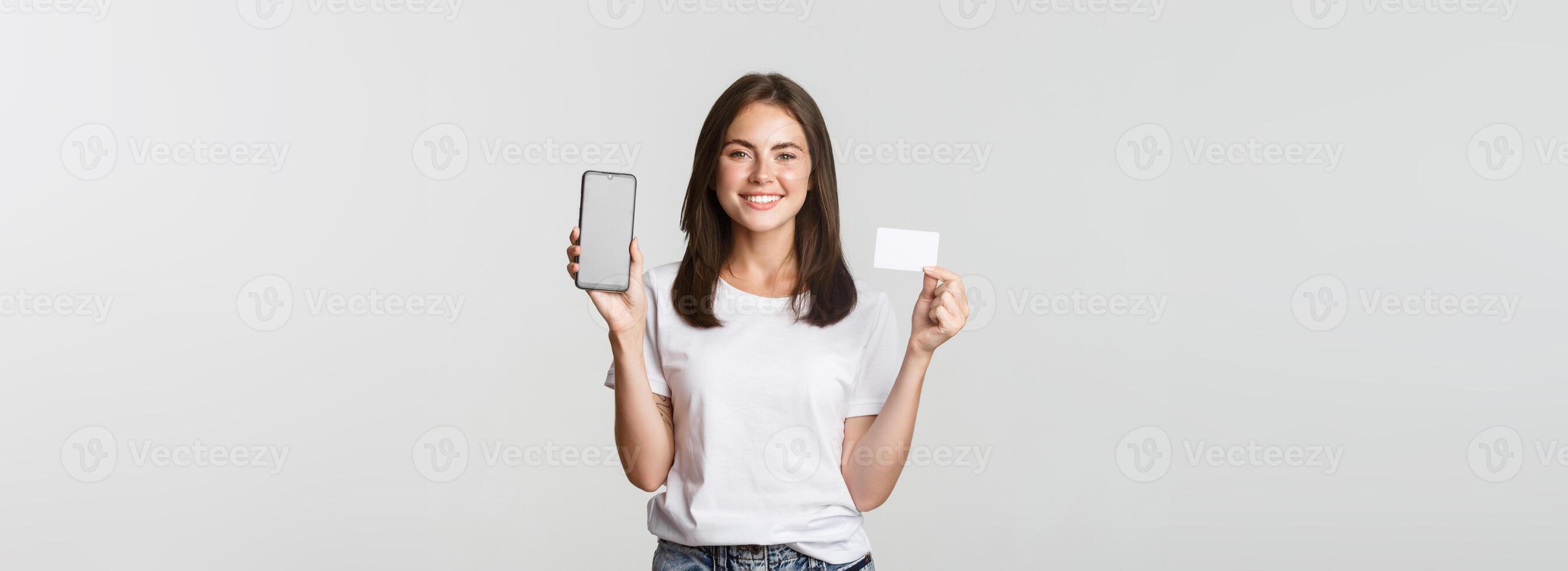Attractive smiling girl looking satisfied and showing credit card, mobile phone screen photo