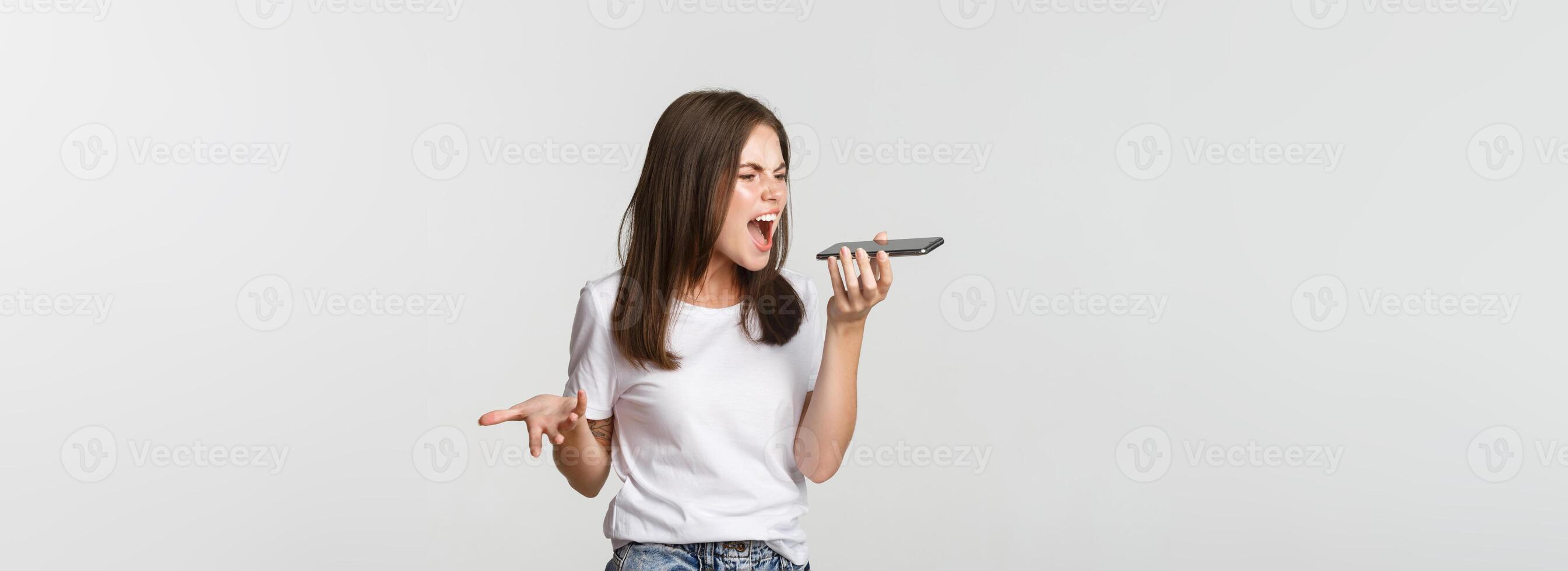 Angry, pissed-off brunette girl arguing, record voice message on mobile phone, white background photo