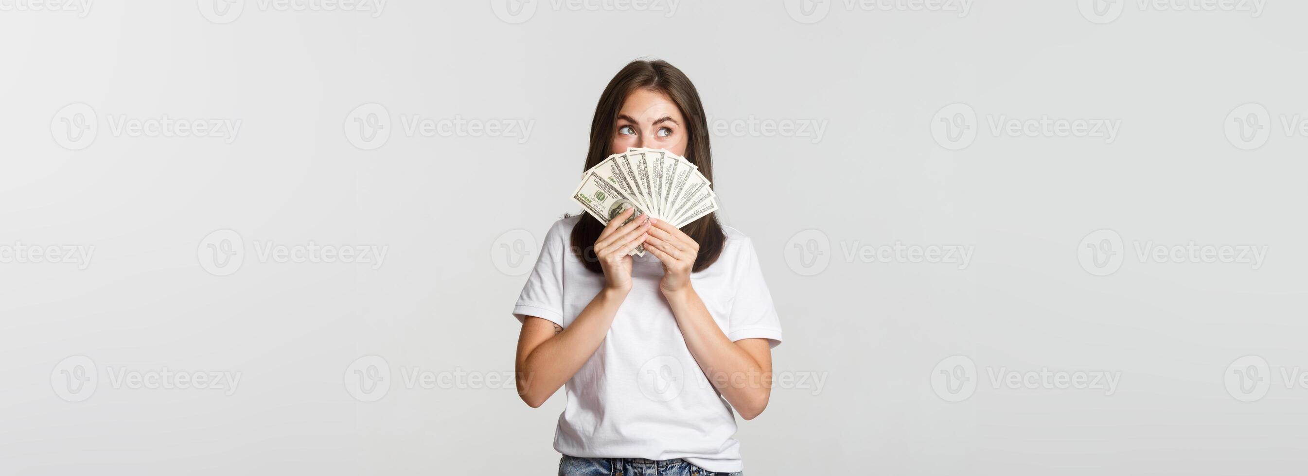 Excited dreamy girl holding money and looking upper left corner, thinking over white background photo