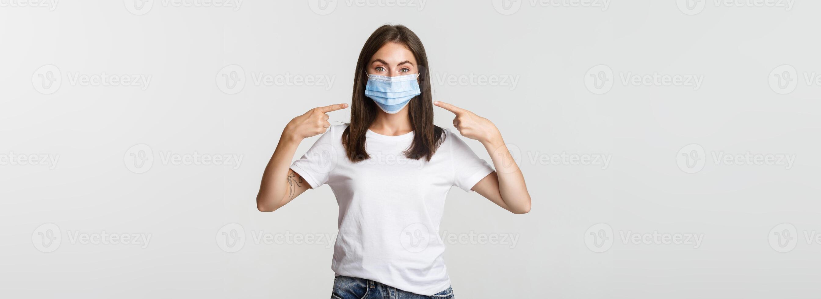 Covid-19, health and social distancing concept. Attractive brunette girl in medical mask pointing finger at face, white background photo