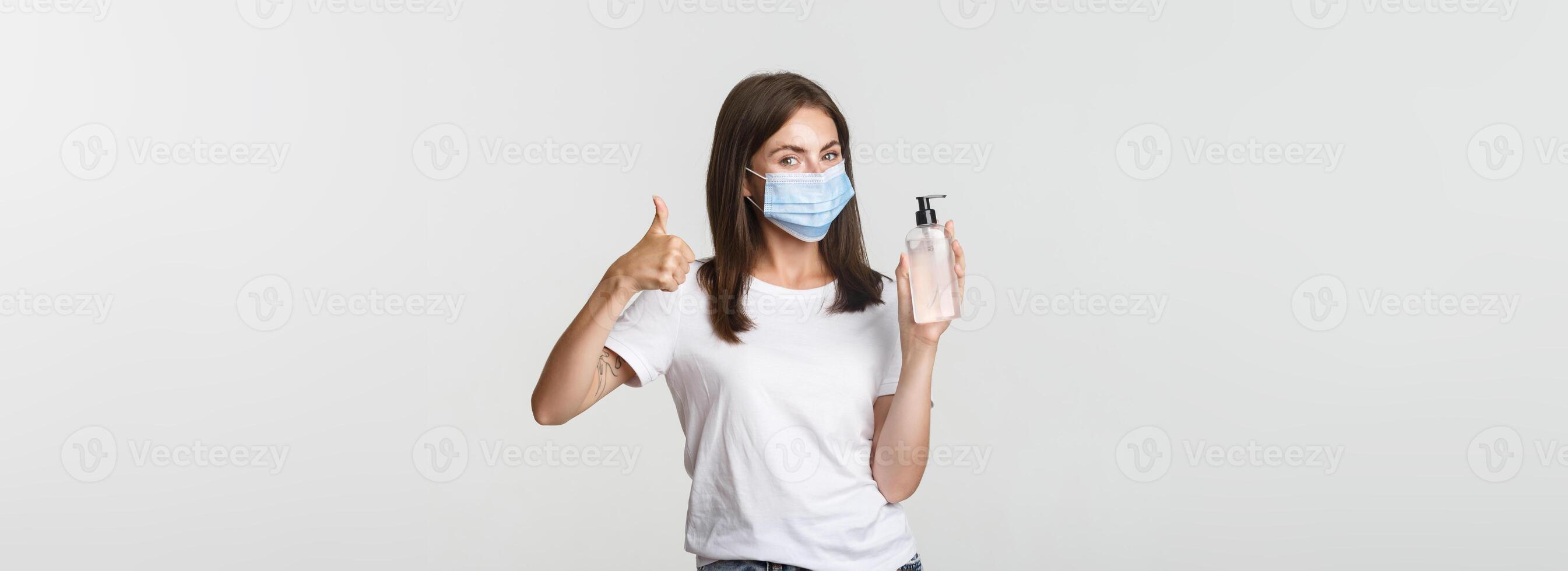 Covid-19, health and social distancing concept. Portrait of satisfied smiling young girl in medical mask, showing thumbs-up and hand sanitizer photo