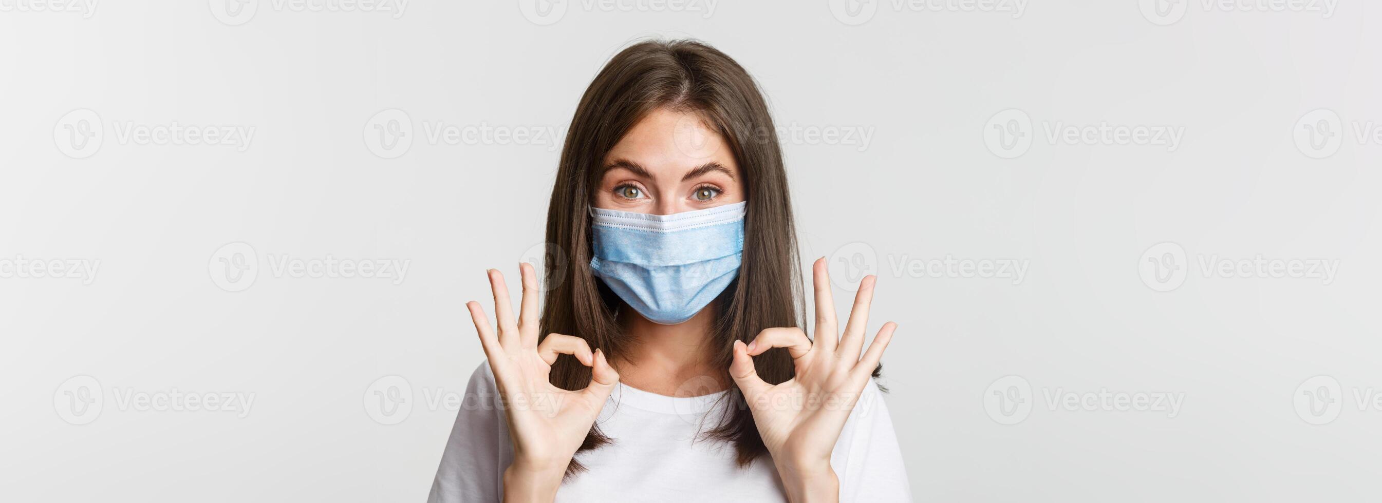 Covid-19, health and social distancing concept. Close-up of pretty satisfied girl in medical mask showing okay gesture in approval photo