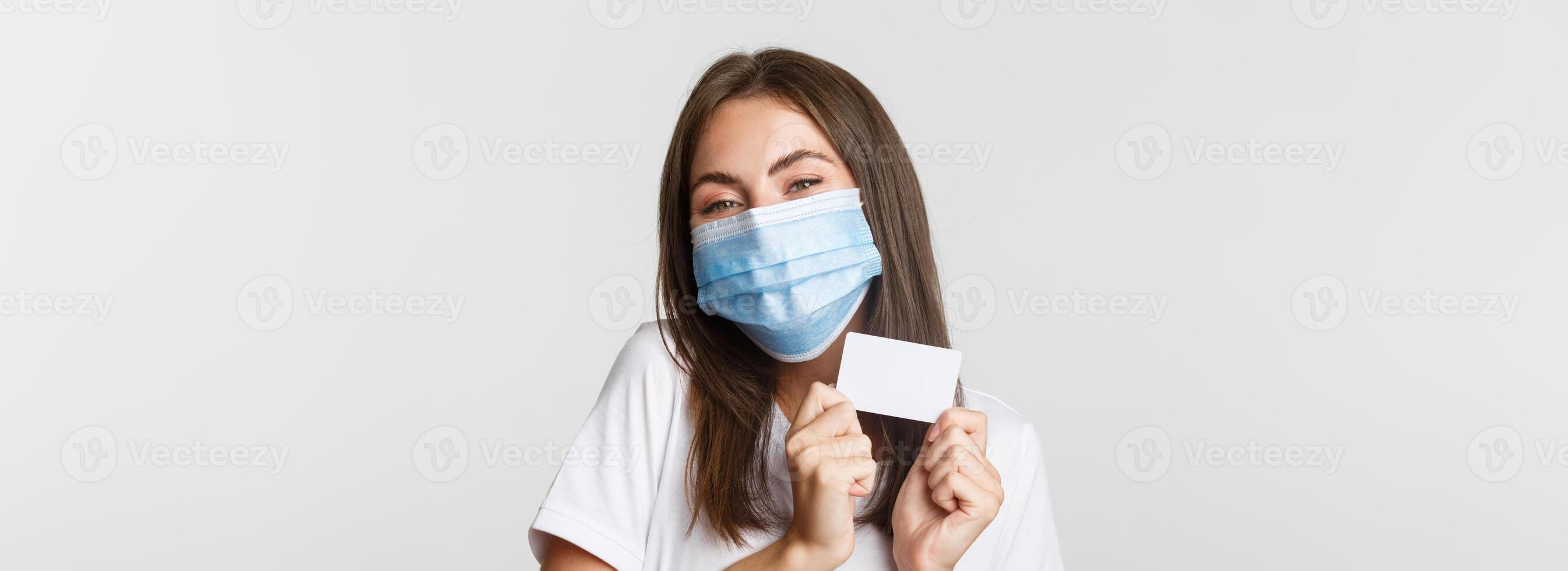 Covid-19, health and social distancing concept. Close-up of attractive happy brunette girl in medical mask showing credit card photo