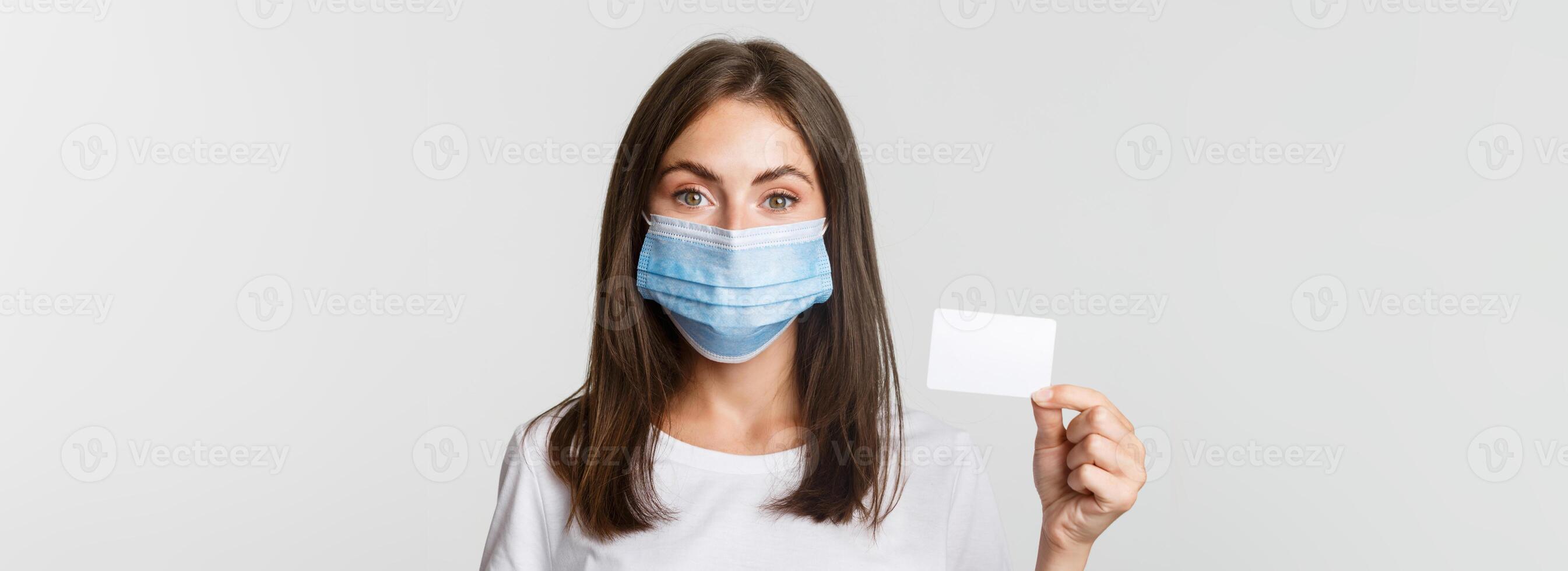 Covid-19, health and social distancing concept. Close-up of pretty brunette girl in medical mask, showing credit card photo