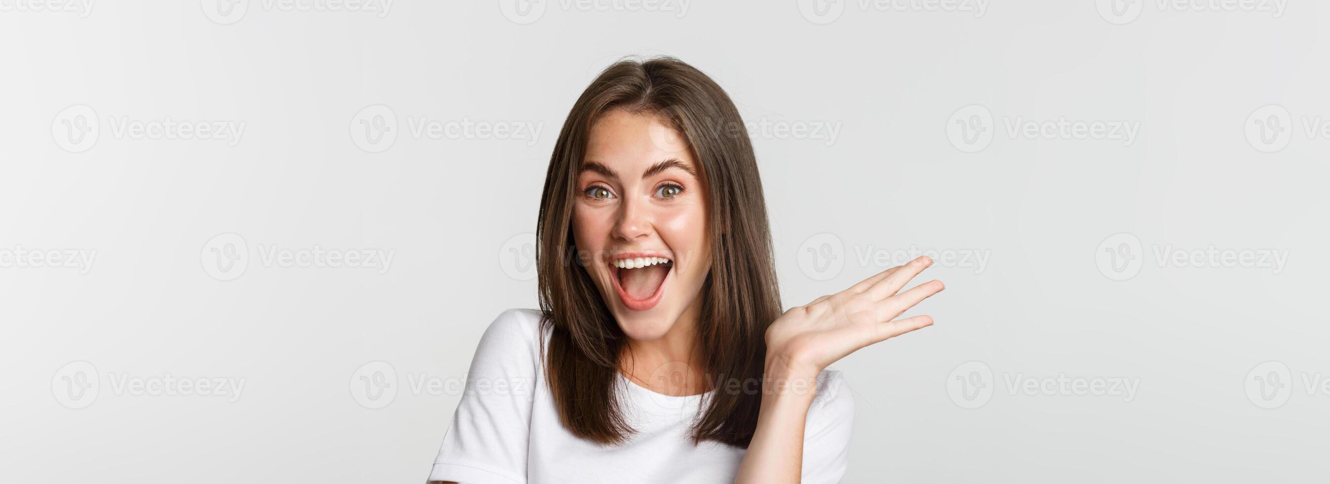 Close-up of excited beautiful brunette girl looking amused, waving hand to say hello, white background photo
