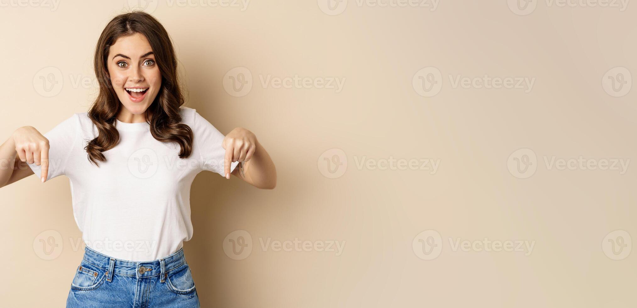 Smiling happy girl pointing fingers down and showing banner, company logo below, click link, standing over beige background photo