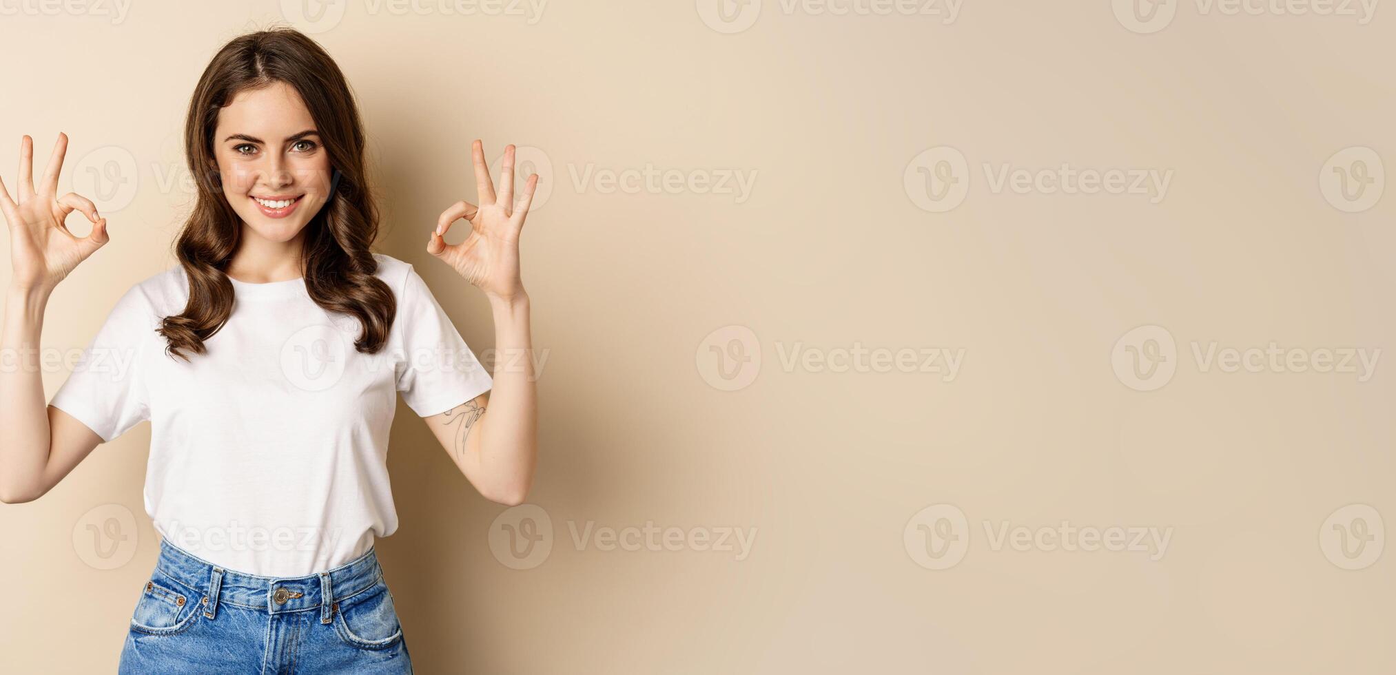 Beautiful female model smiling pleased, showing okay, zero sign, complimenting, praising you, standing over beige background photo