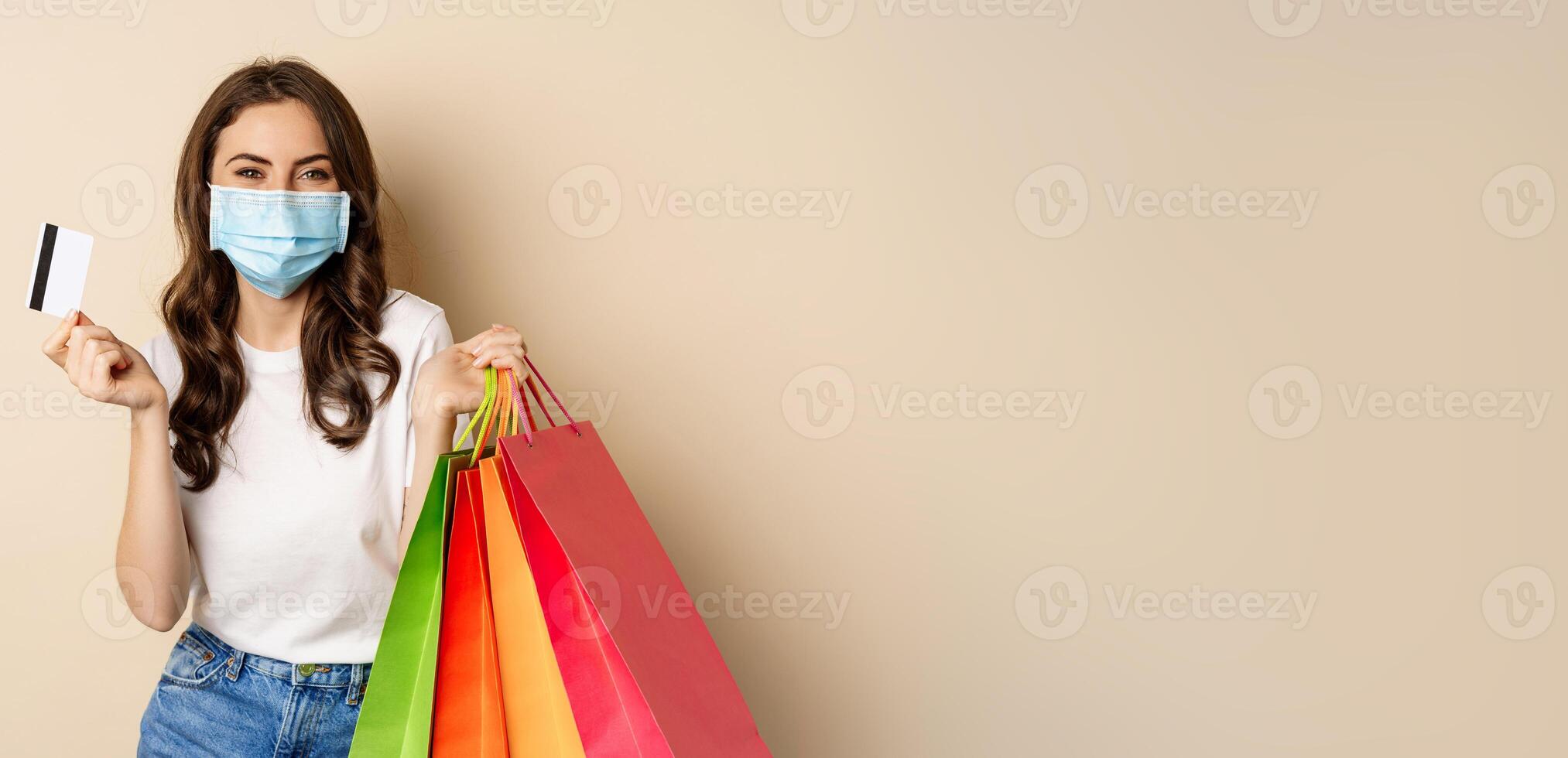 Covid-19 pandemic and lifestyle concept. Young woman posing in medical face mask with shopping bags from mall, holding credt card in hand, beige background photo