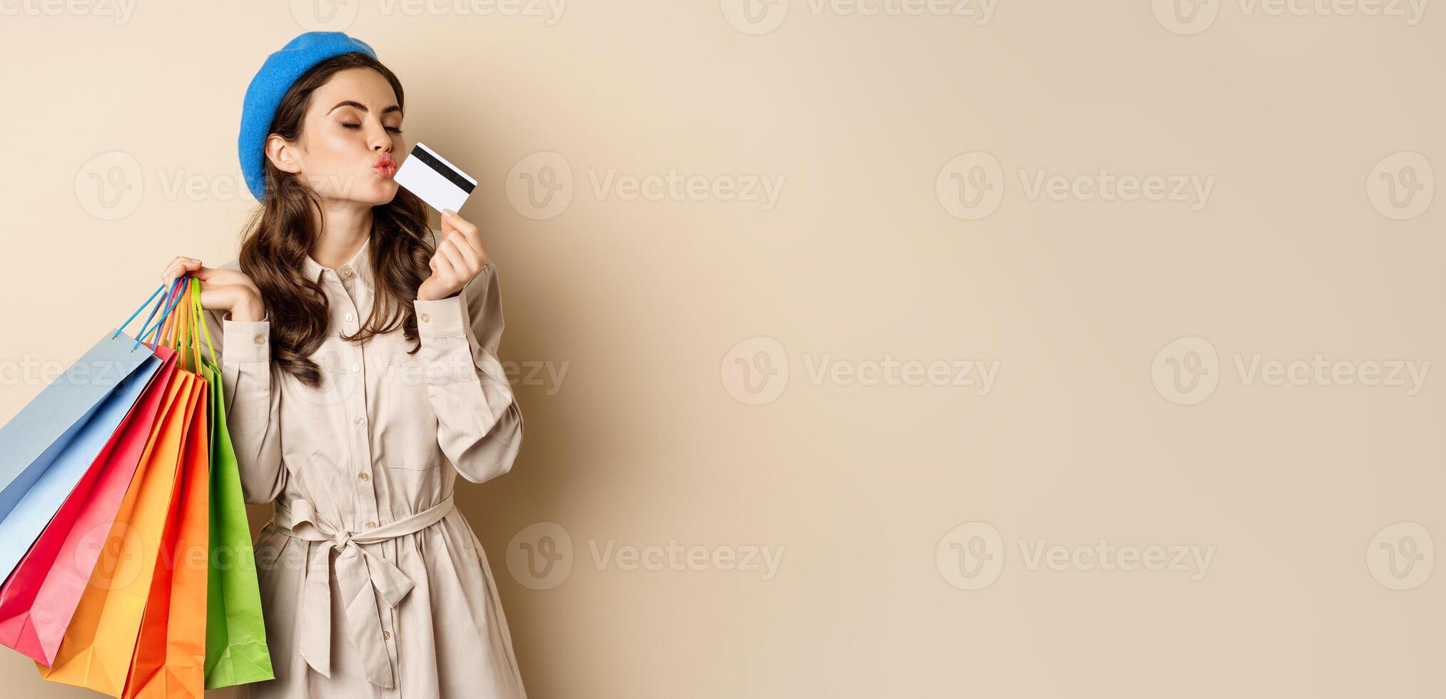 Portrait of cute happy girl kissing her credit card after buying gifts, holding shopping bags, standing over beige background photo