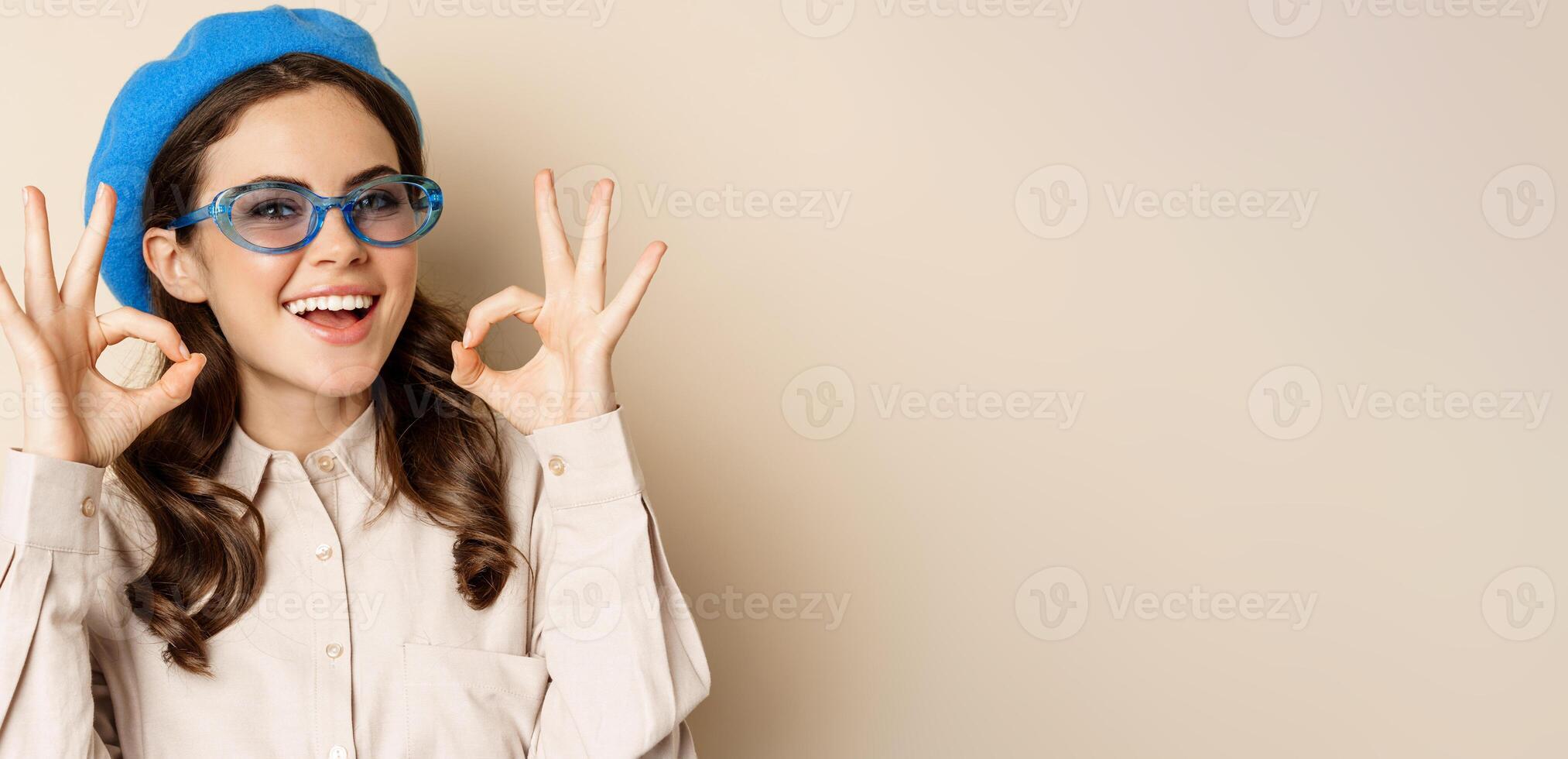 Image of happy smiling woman in stylish sunglasses, showing okay signs in approval, recommending eyewear store, beige background photo