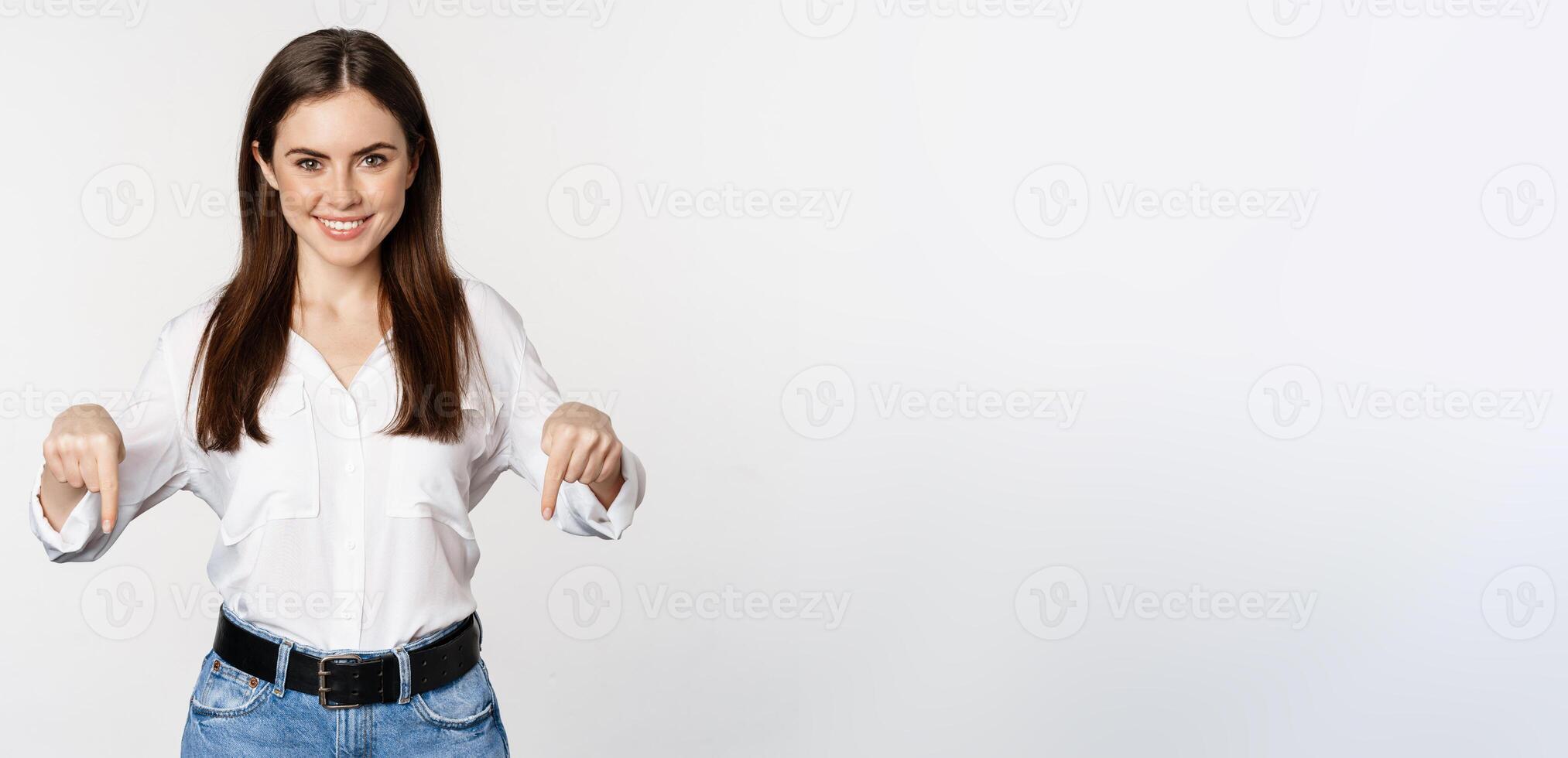 Confident smiling woman pointing fingers down, showing logo, banner below, recommending click on link, standing over white background photo