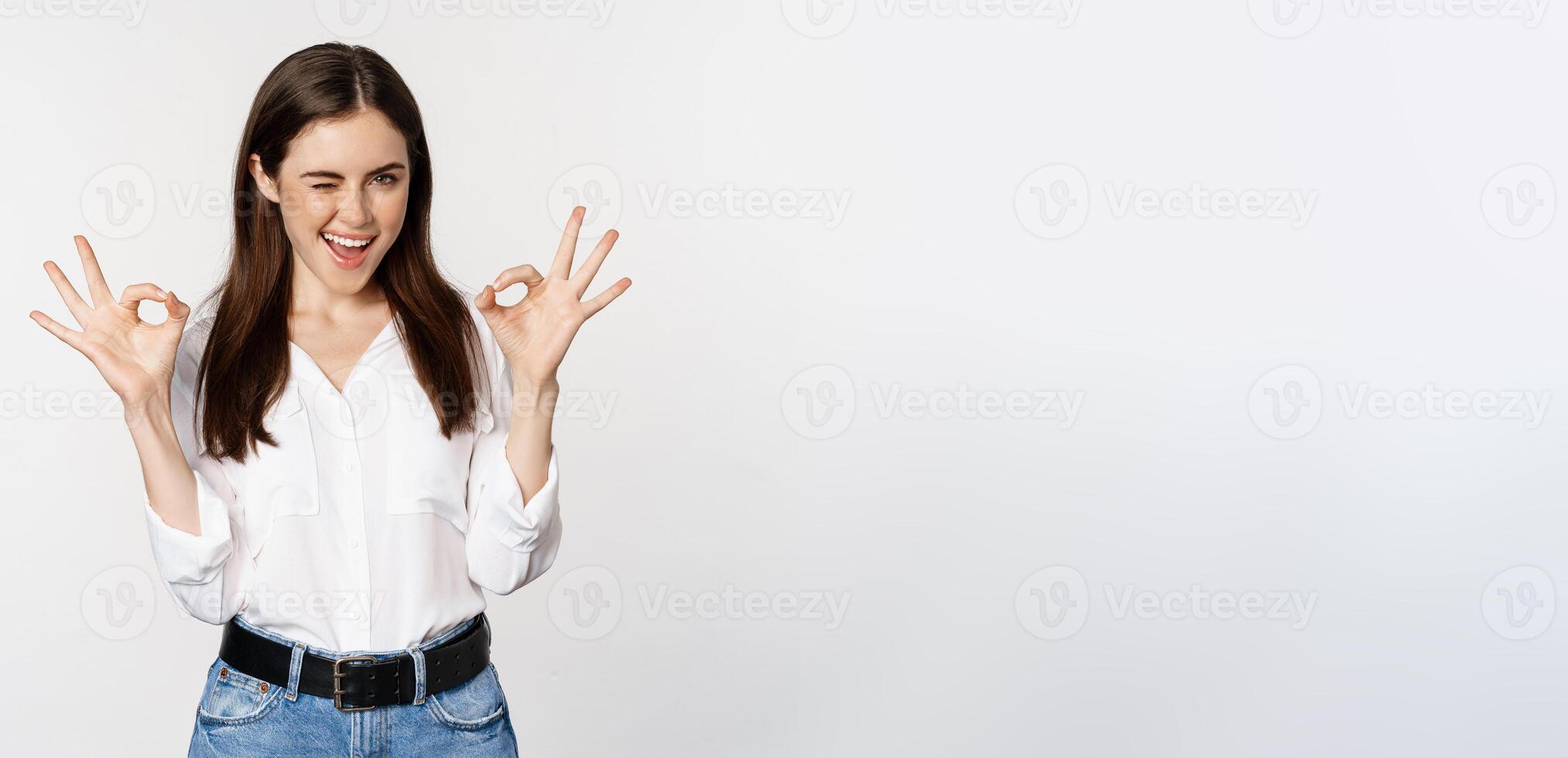 Smiling confident business woman, winking, showing okay ok sign, excellent, no problem gesture, standing over white background photo