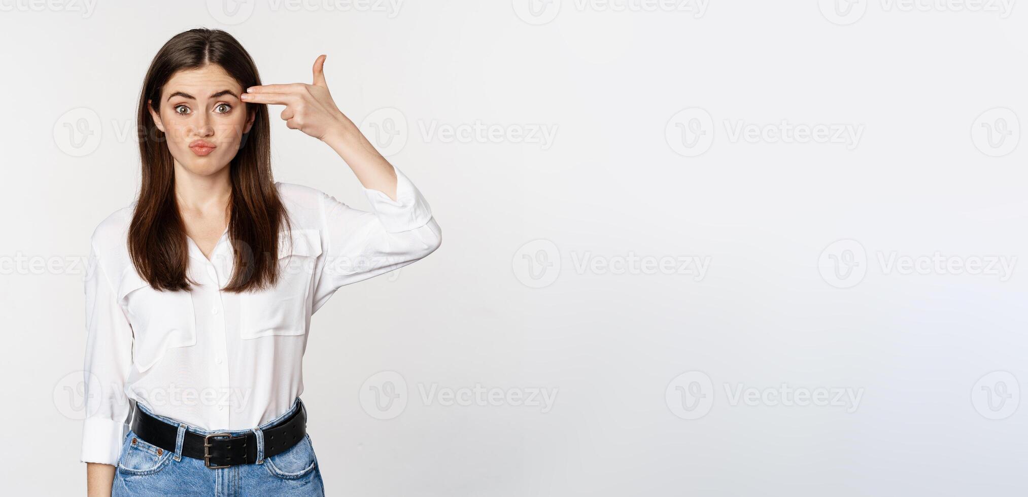 Image of annoyed young woman showing finger gun gesture over head, bothered by smth, tired, standing over white background photo