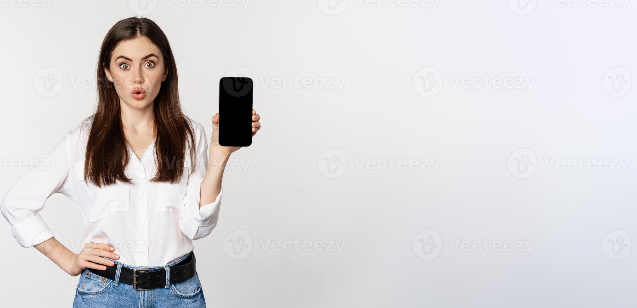 Enthusiastic corporate woman demonstrating website, mobile phone screen, showing application and saying wow, standing over white background photo
