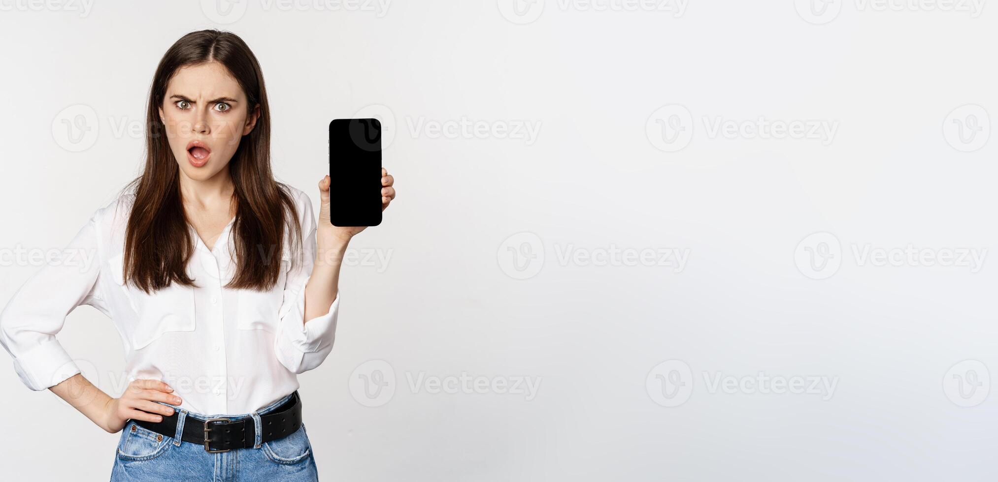 Shocked and frustrated woman showing mobile phone screen app, smartphone application, looking insulted, standing over white background photo