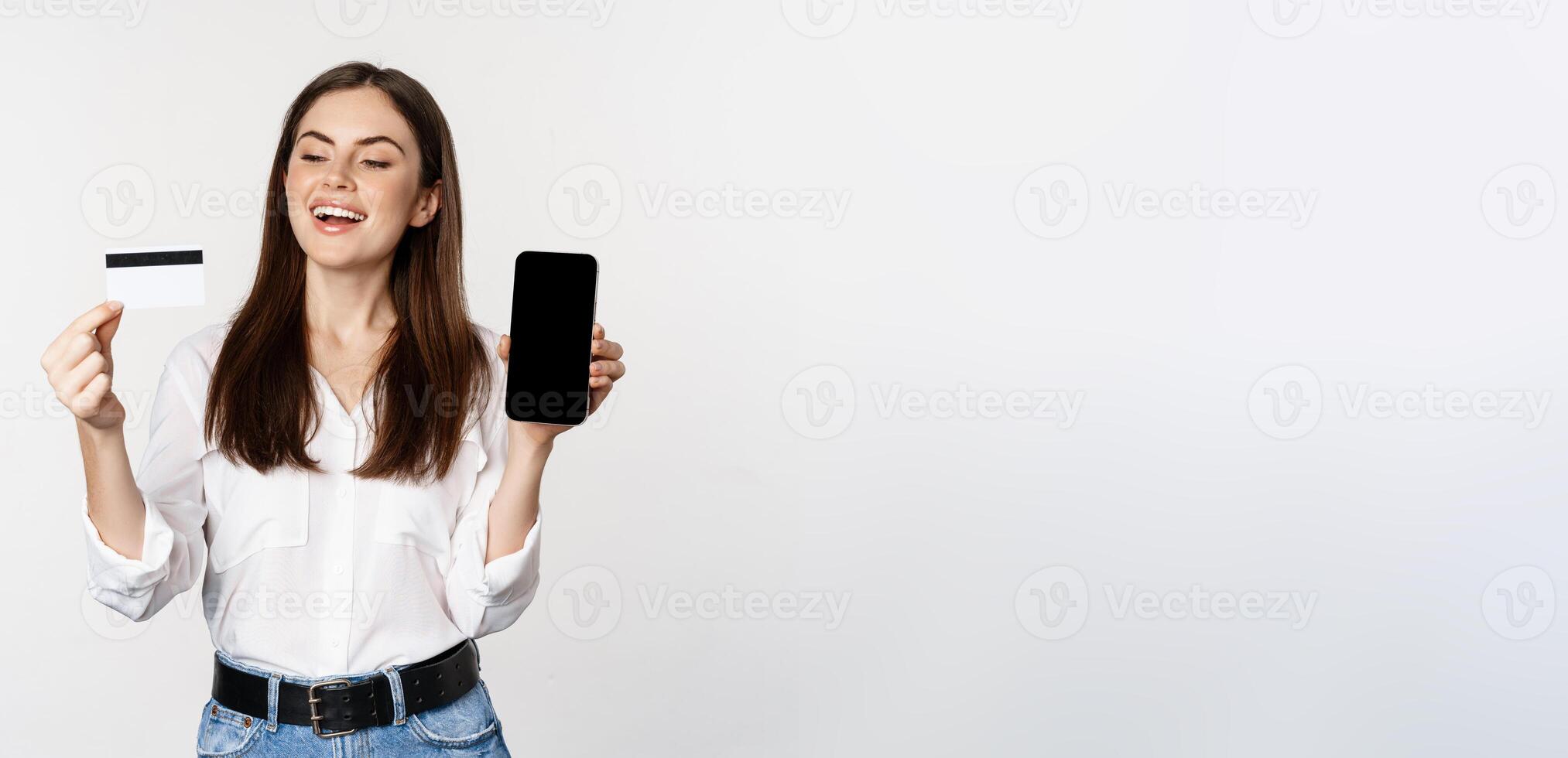 Happy woman showing credit card and smartphone screen, concept of online shopping, buying in app, standing over white background photo