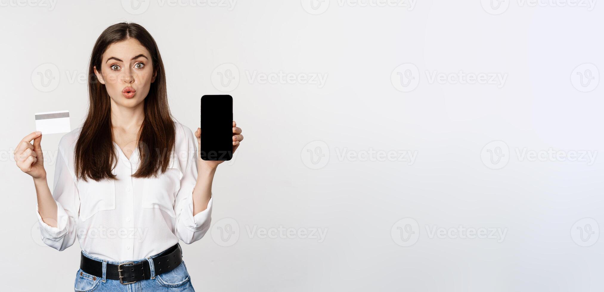 Happy woman showing credit card and smartphone screen, concept of online shopping, buying in app, standing over white background photo
