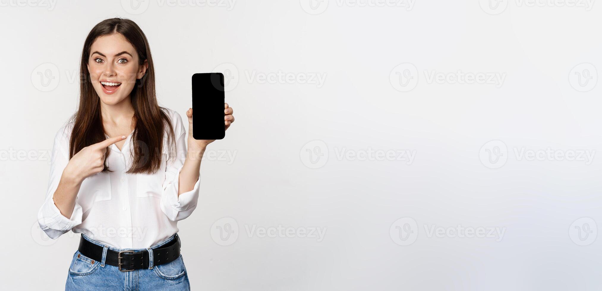 Happy beautiful woman smiling, pointing at mobile phone screen, showing advertisement, website, standing over white background photo