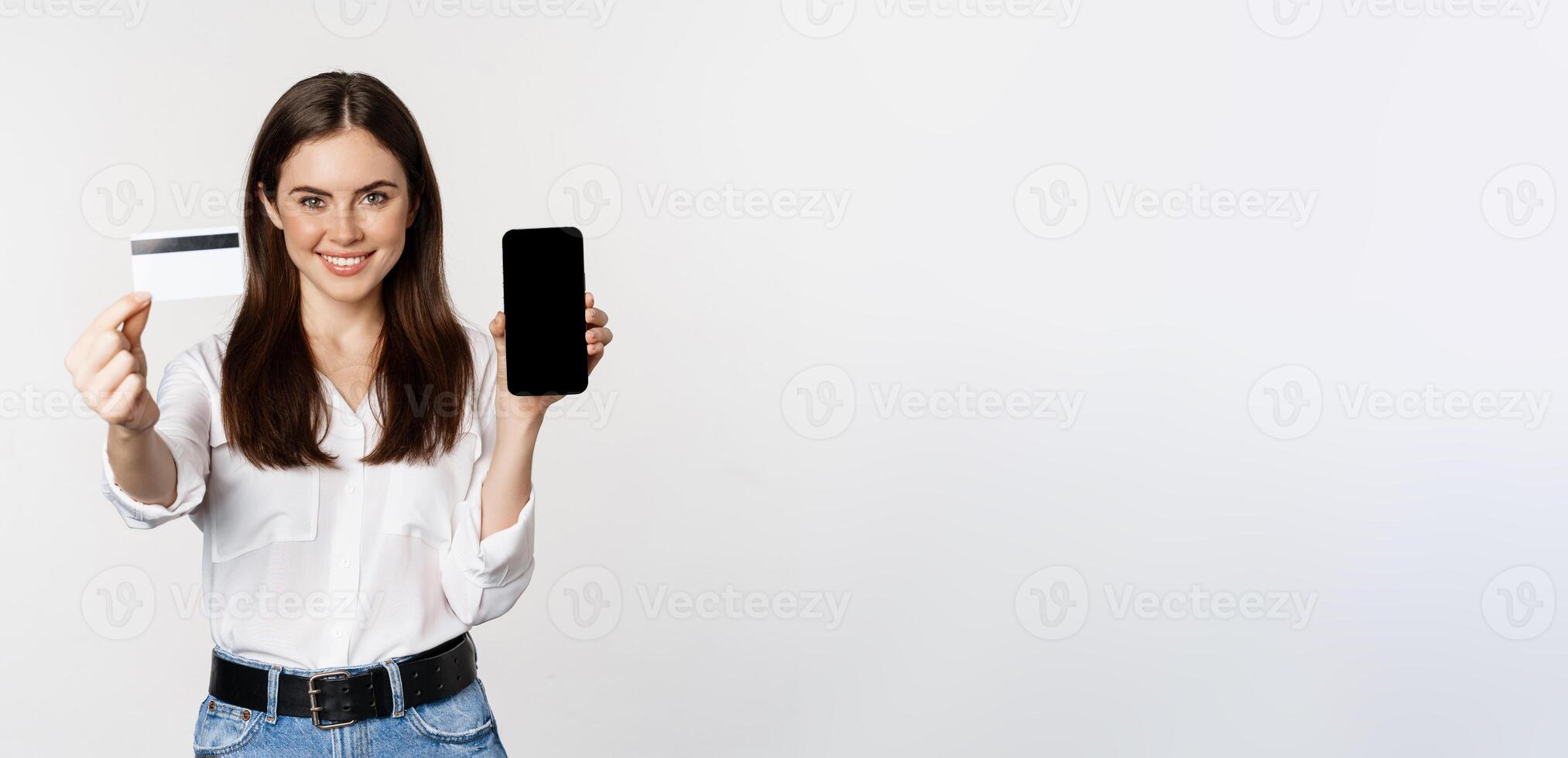 Portrait of female model showing credit card with smartphone screen, recommending application, standing over white background photo
