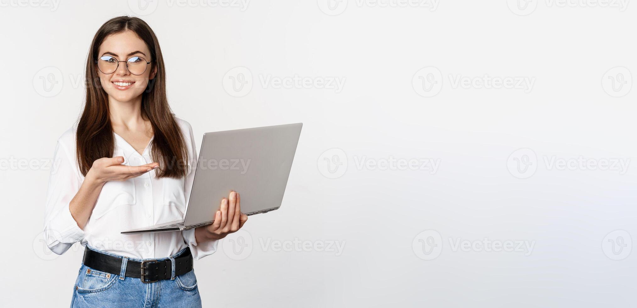 Portrait of woman in glasses holding laptop, pointing at screen, showing her work on computer, standing over white background photo
