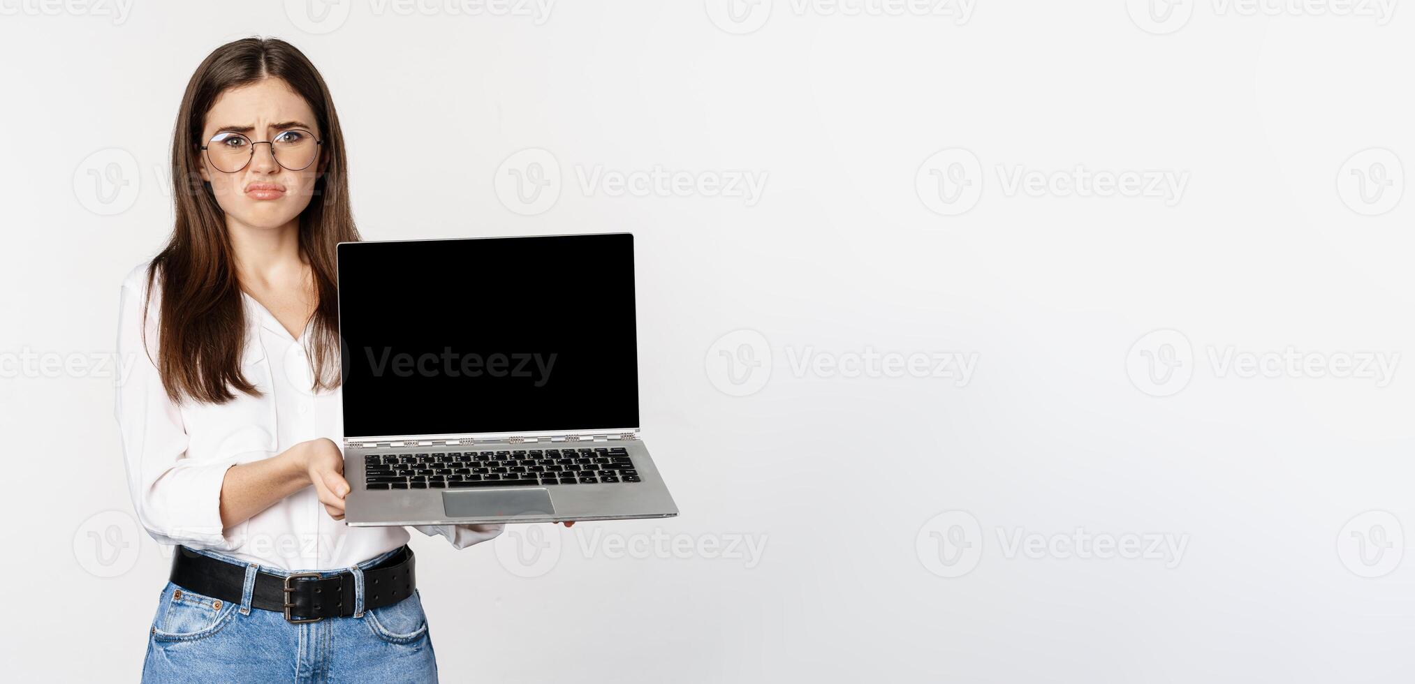 Sad brunette girl, student showing laptop screen and grimacing upset, standing disappointed against white background photo