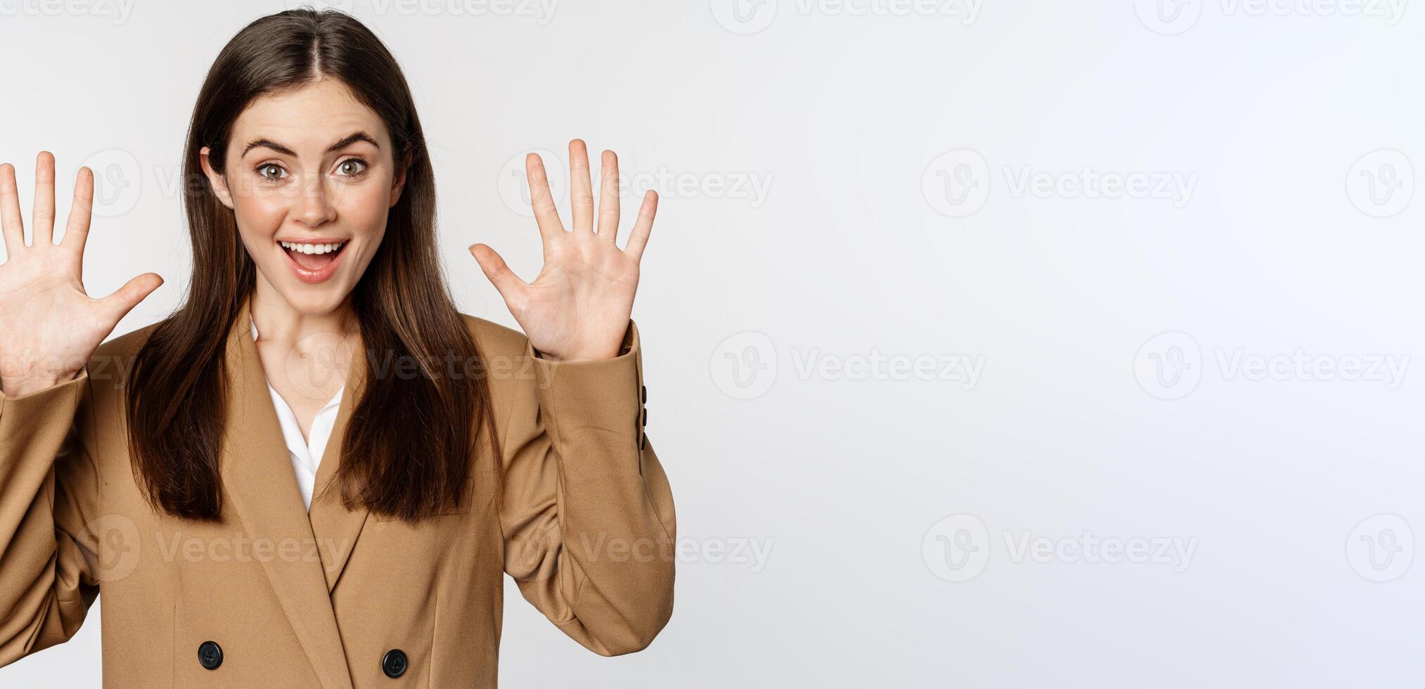 Enthusiastic businesswoman in suit, raising hands palms up, number ten dozen gesture and smiling, standing over white background photo