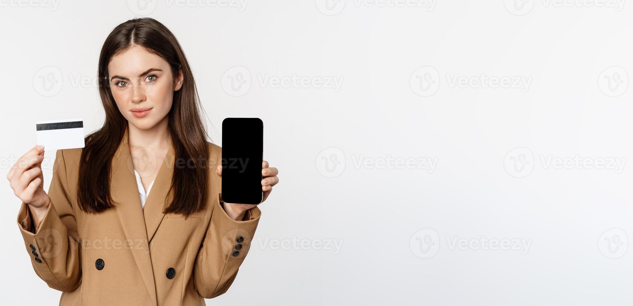 Businesswoman showing mobile phone screen and credit card, demonstrating application on smartphone, standing in suit over white background photo
