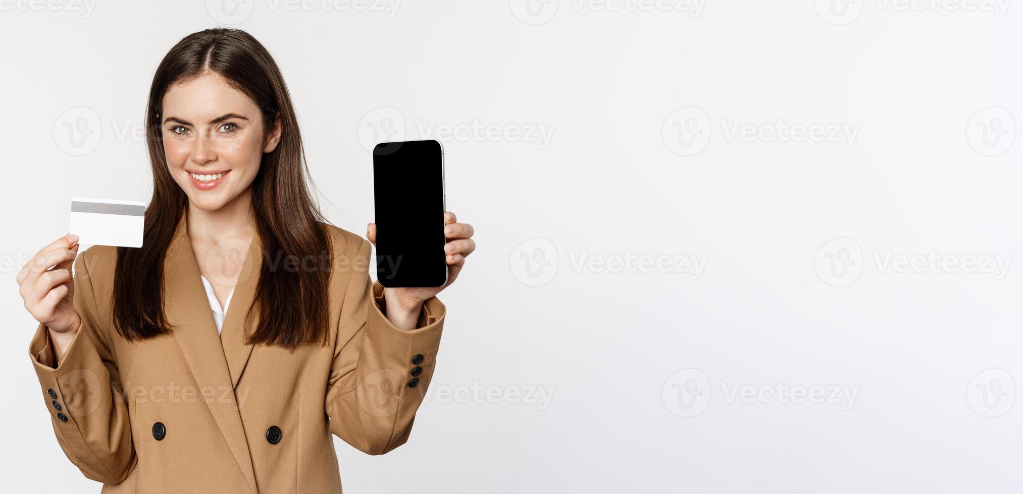 Businesswoman showing mobile phone screen and credit card, demonstrating application on smartphone, standing in suit over white background photo