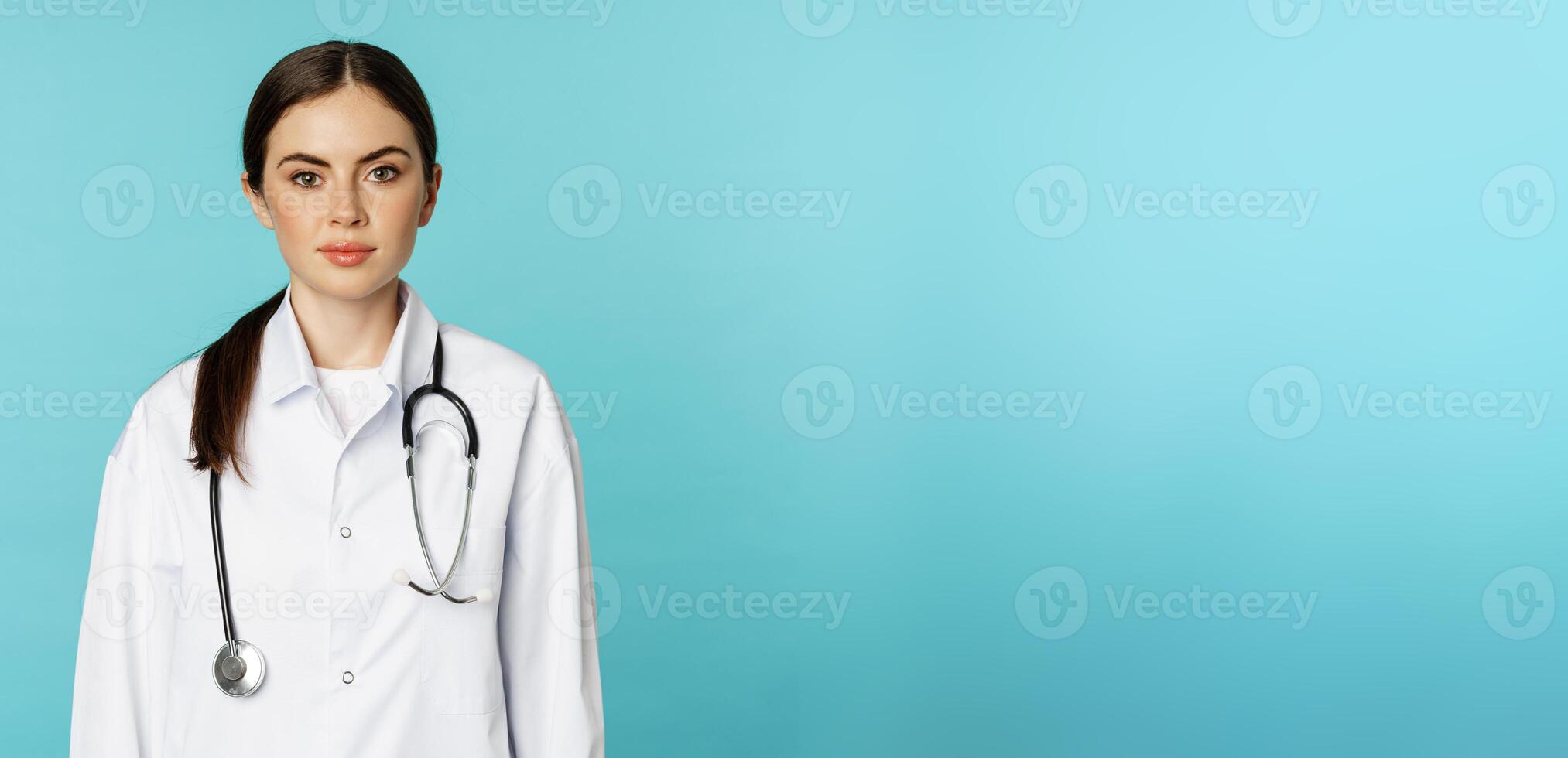 Medical staff and doctors concept. Young smiling female doctor, healthcare worker in white coat and stethoscope, looking confident, waiting patients, blue background photo