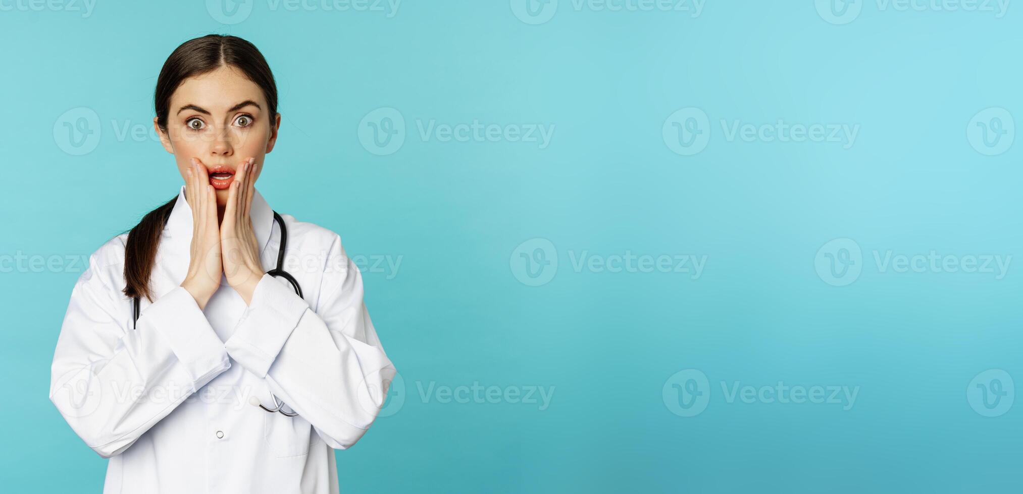 Portrait of shocked woman doctor, female hospital intern in white coat, looking concerned and confused at camera, disbelief face, standing over torquoise background photo