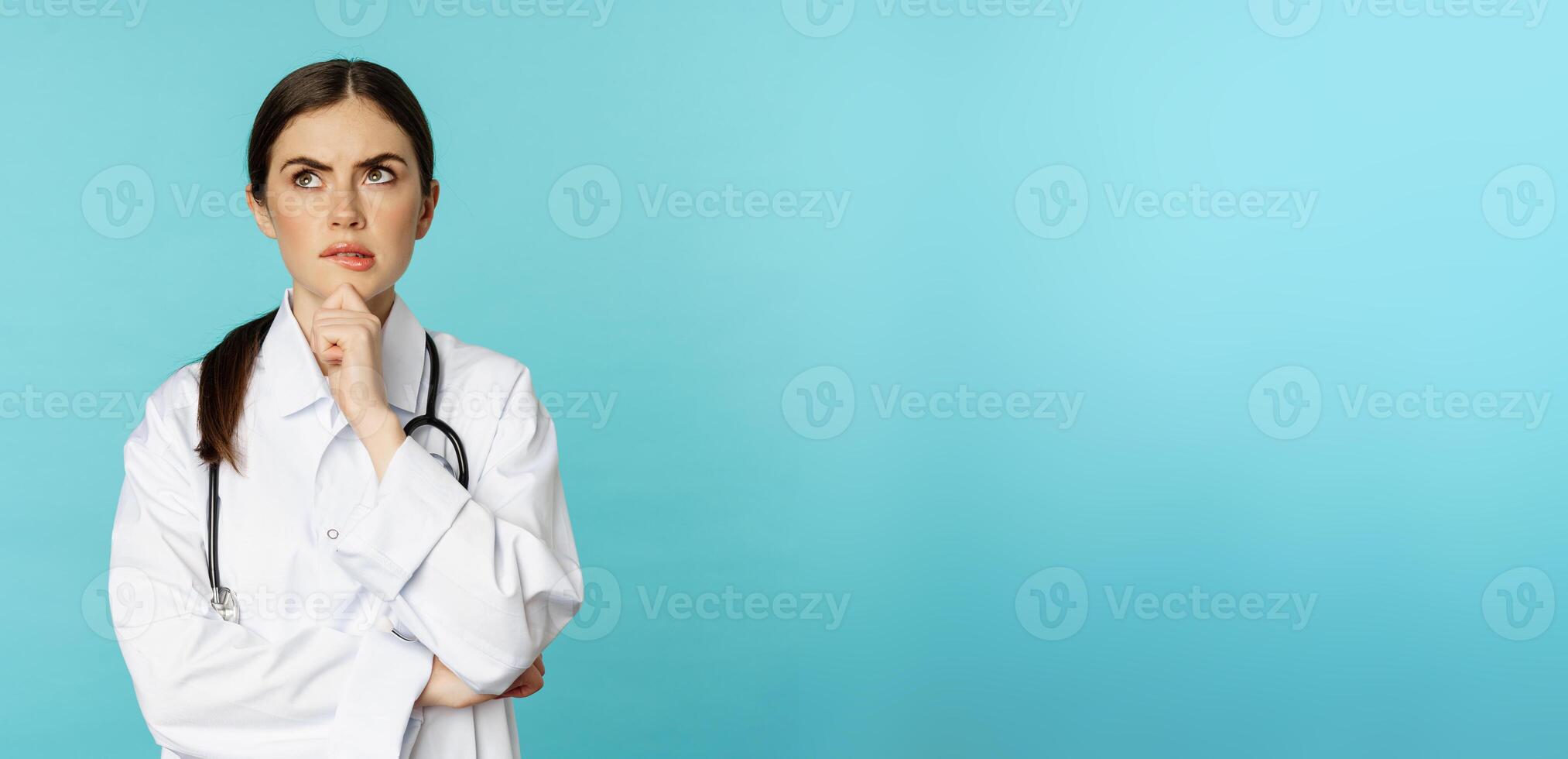 Young female doctor, hospital worker in white coat, thinking and looking away thoughtful, searching solution, standing over toquoise background photo